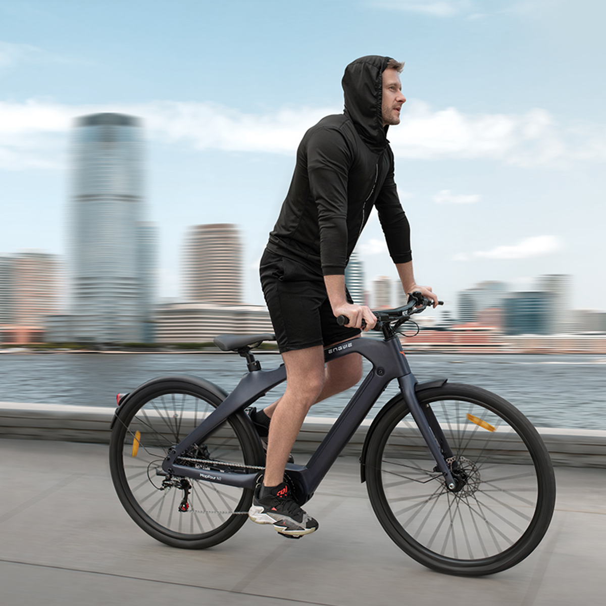 Mężczyzna jadący ciemnoszarym rowerem elektrycznym. Ma na sobie czarny strój i jedzie w pobliżu panoramy miasta.