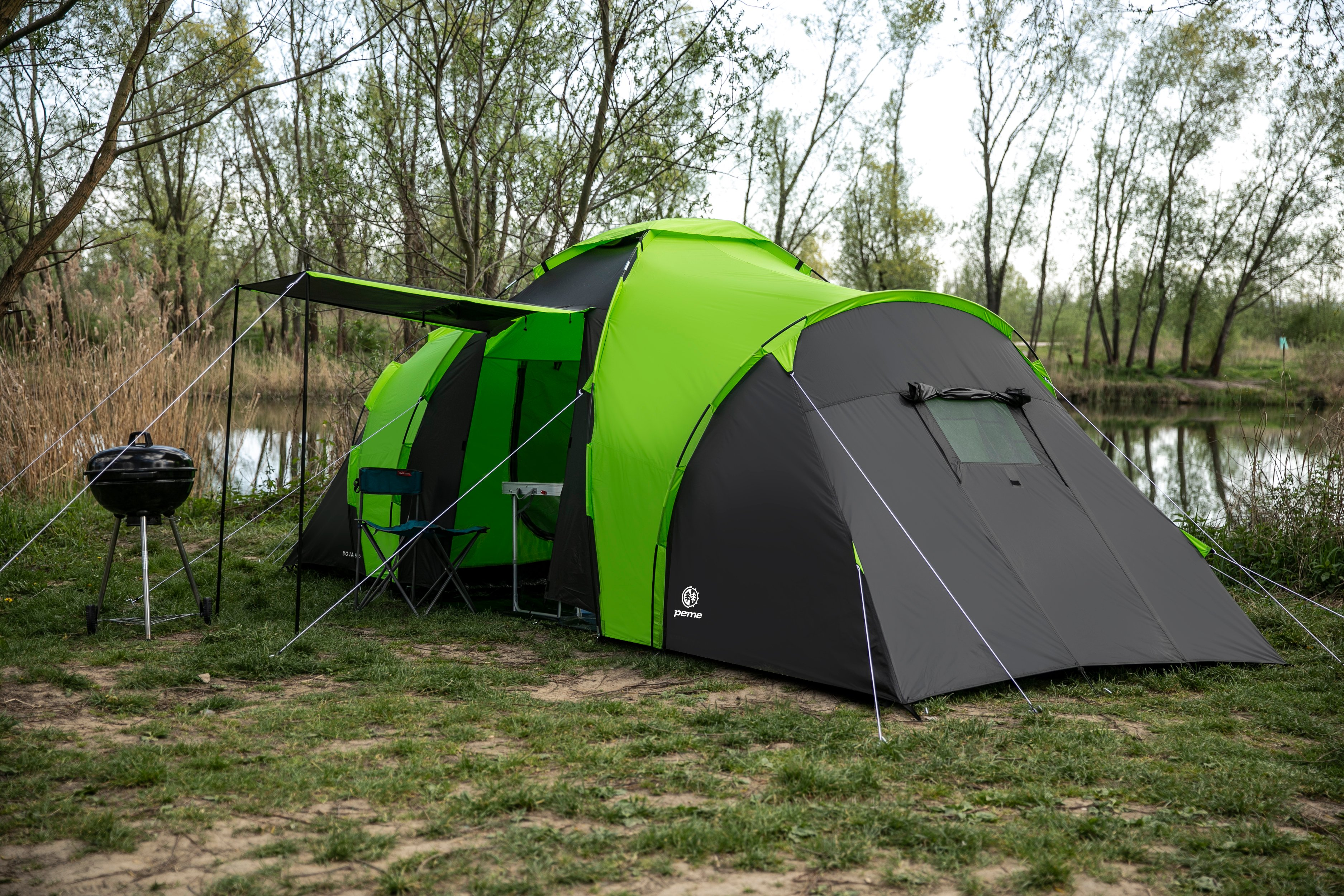 Grün-schwarzes Zelt, Grill, Tisch und Stuhl auf Gras neben See, Bäume im Hintergrund.
