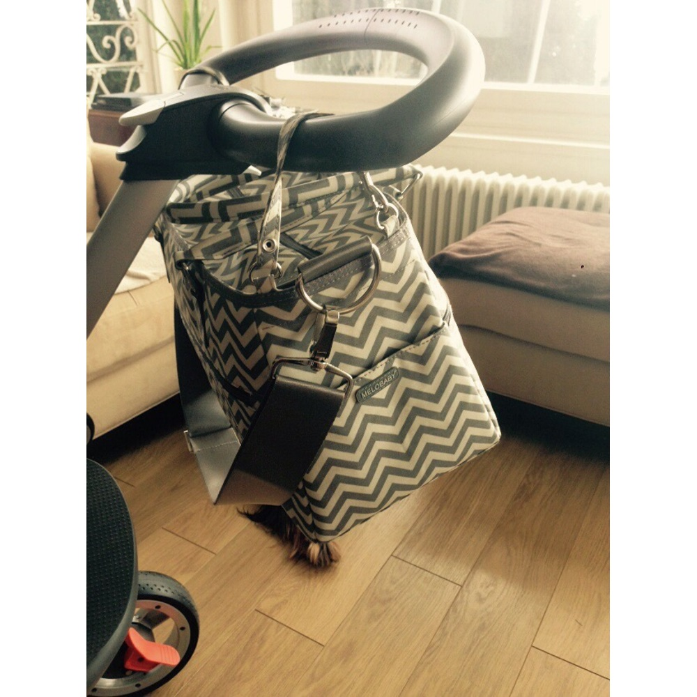 Ein Kinderwagen mit einer daran hängenden Wickeltasche, an einem Fenster. Die Tasche hat ein Zickzackmuster.