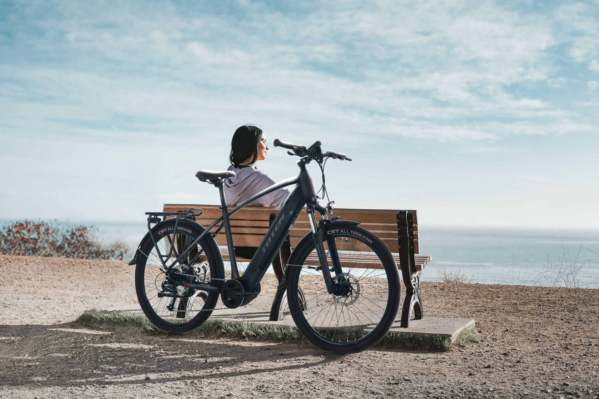 Eine Frau sitzt auf einer Bank neben einem E-Bike und blickt unter blauem Himmel aufs Meer.