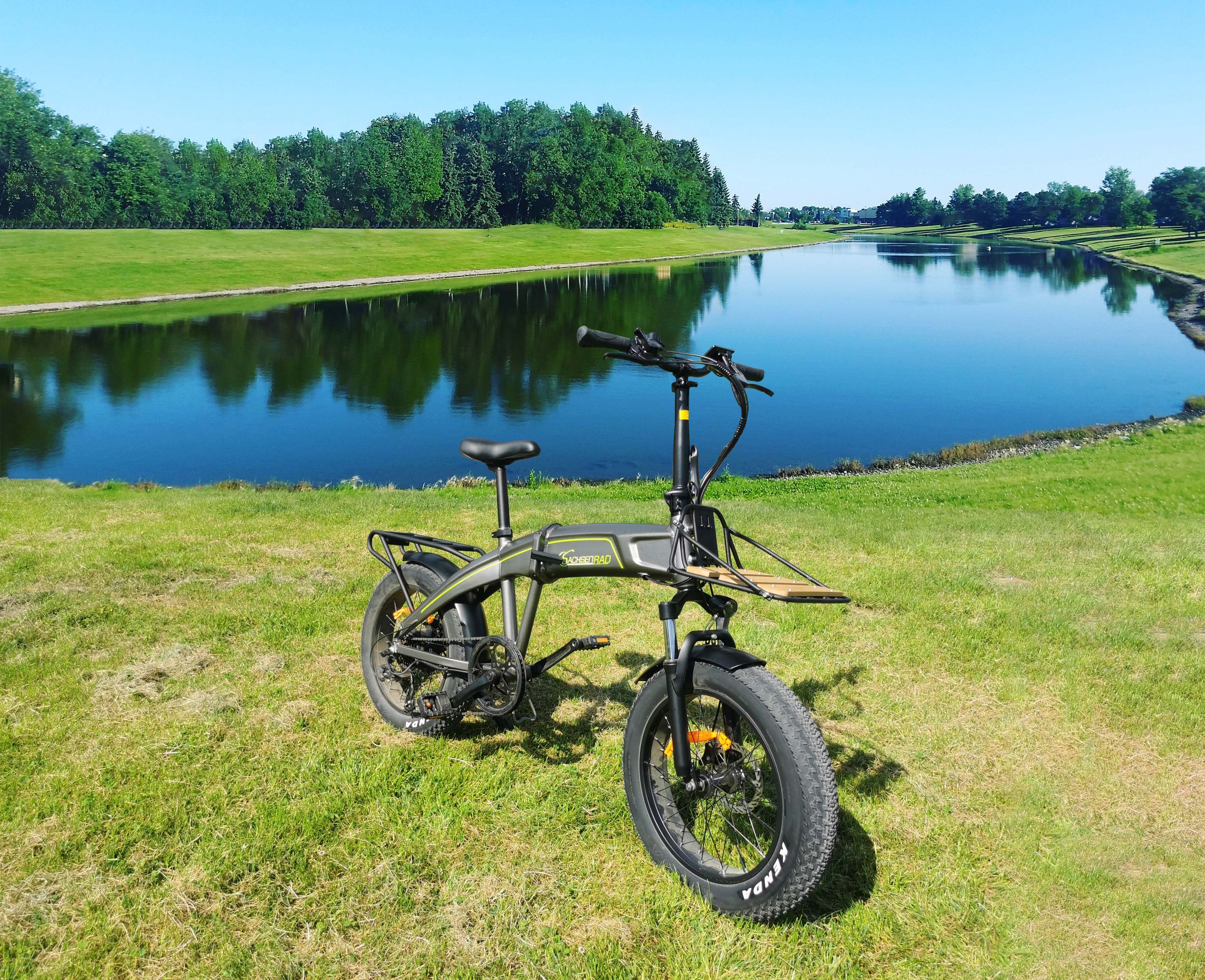 Elektrofahrrad auf Gras am See. Grüne Bäume im Hintergrund und blauer Himmel.