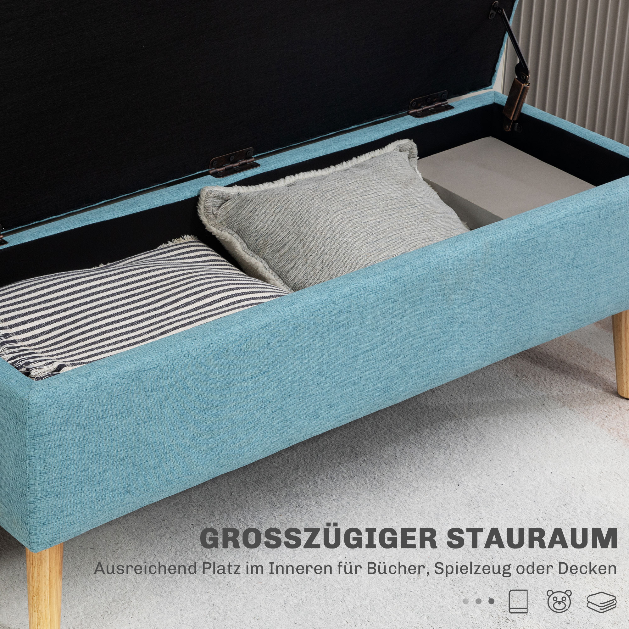 Blauer Stauraumbank mit Kissen. Helle Holzbeine, weißer Boden. Offener Deckel zeigt den Stauraum.