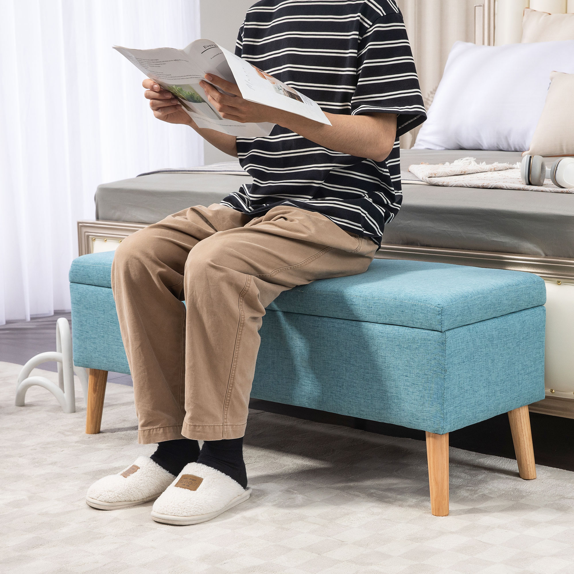 Mann liest auf blauer Bank mit Holzbeinen. Er trägt gestreiftes Hemd und Hose. Bett im Hintergrund.