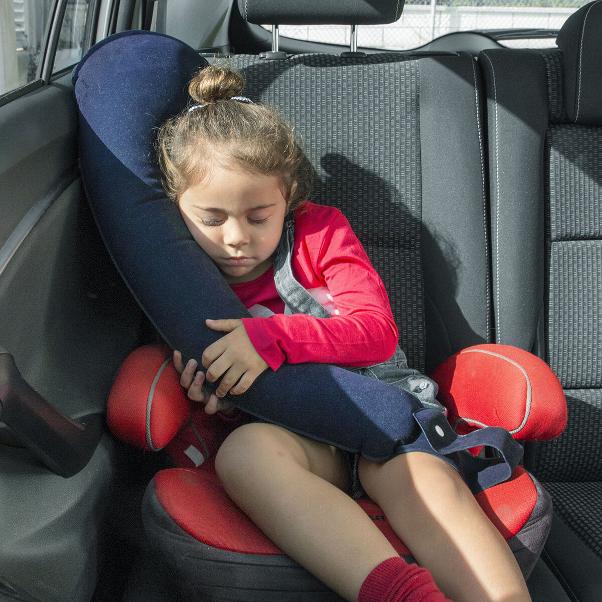 Un bambino che dorme in un seggiolino auto, sostenuto da un cuscino da viaggio blu navy, all'interno di un'auto.