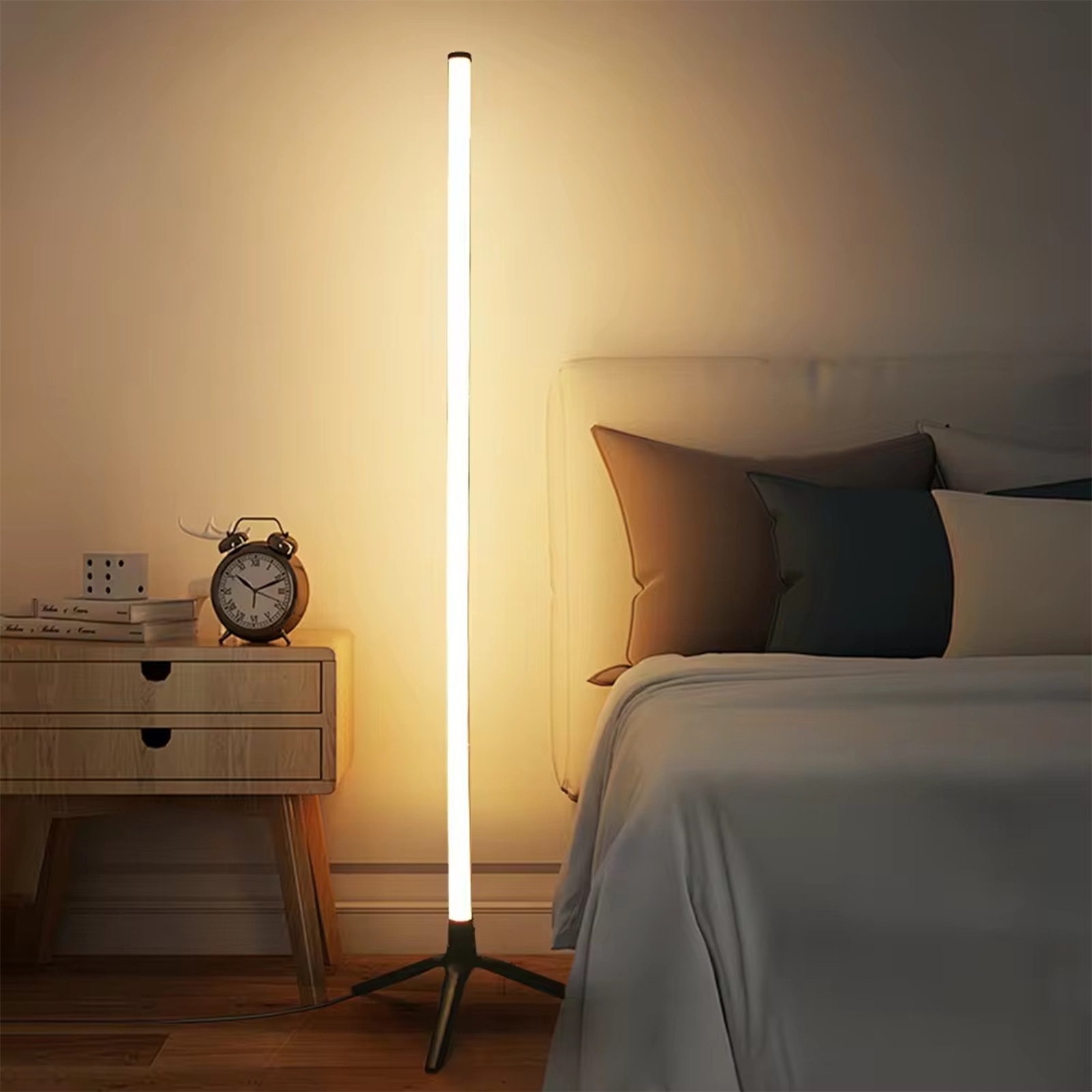 Hohe, leuchtende Stehlampe in einem Schlafzimmer. Steht neben Bett, Kommode und Wecker.