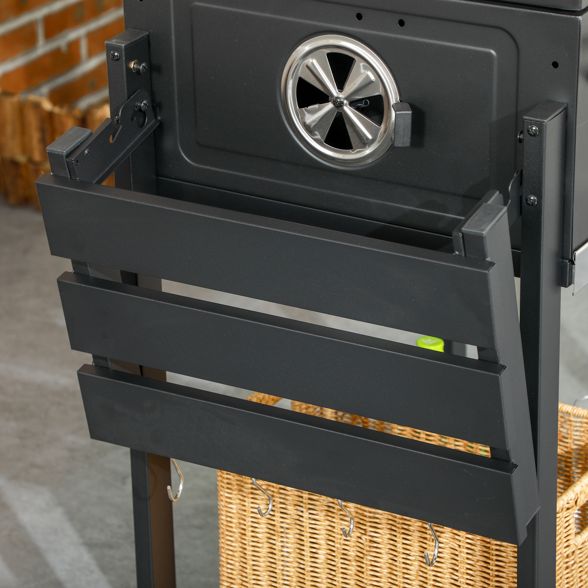 Barbecue-Grill mit Seitenablage. Zeigt eine offene Seitenablage mit Haken und Korb darunter.