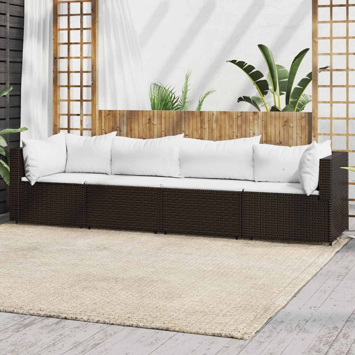 Braunes Rattan-Sofa mit weißen Kissen auf einem beigen Teppich. Holzzaun im Hintergrund, grüne Pflanzen.