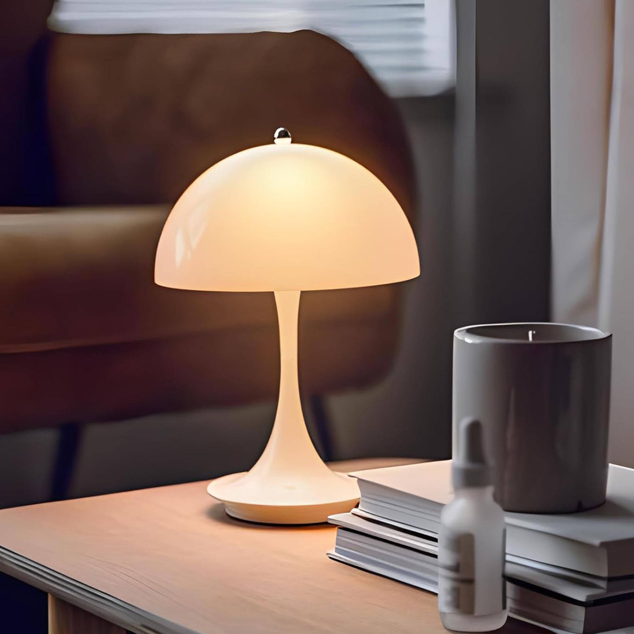 Eine weiße Tischlampe mit pilzförmigem Schirm, auf einem Holztisch mit Büchern, einer Flasche und einer Tasse.