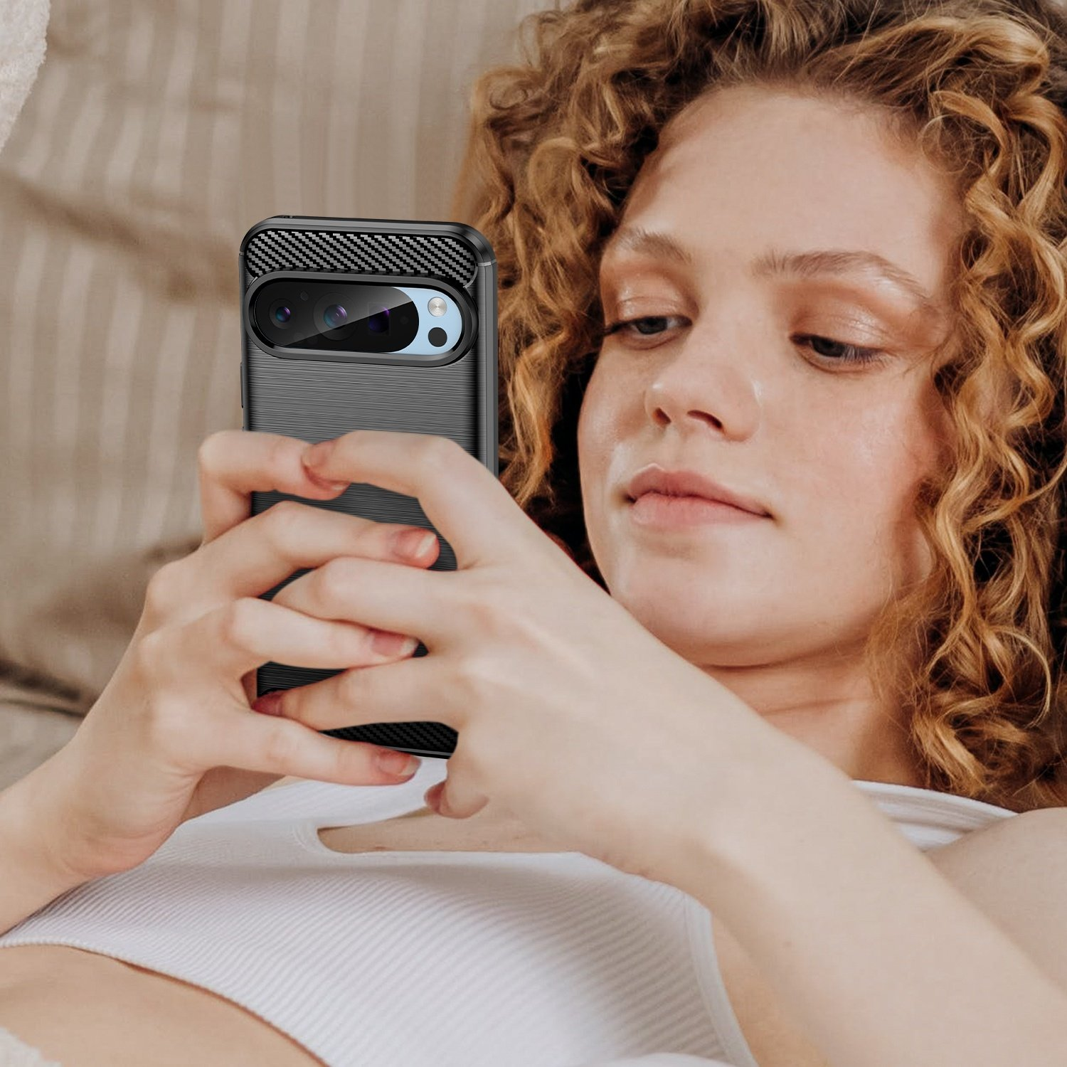 Una mujer sostiene un teléfono con funda de fibra de carbono, mirando la pantalla. Está en la cama.