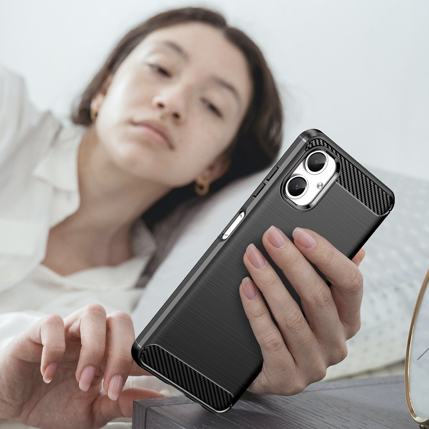 Una mujer sosteniendo un teléfono negro con una funda en sus manos. Está acostada en la cama.
