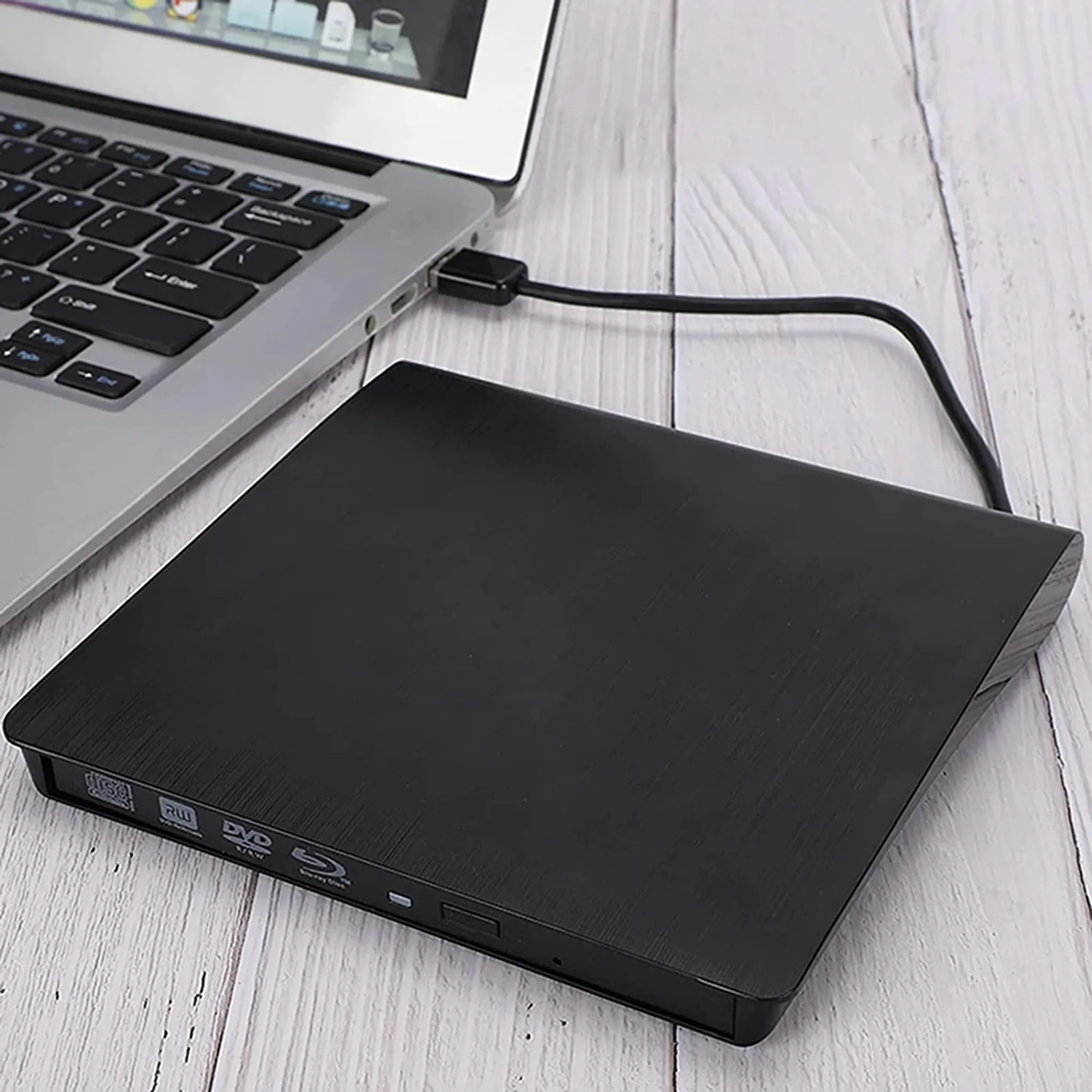 Schwarzes externes DVD-Laufwerk, über USB-Kabel mit Laptop verbunden. Der Laptop ist auf einer Holzoberfläche geöffnet.