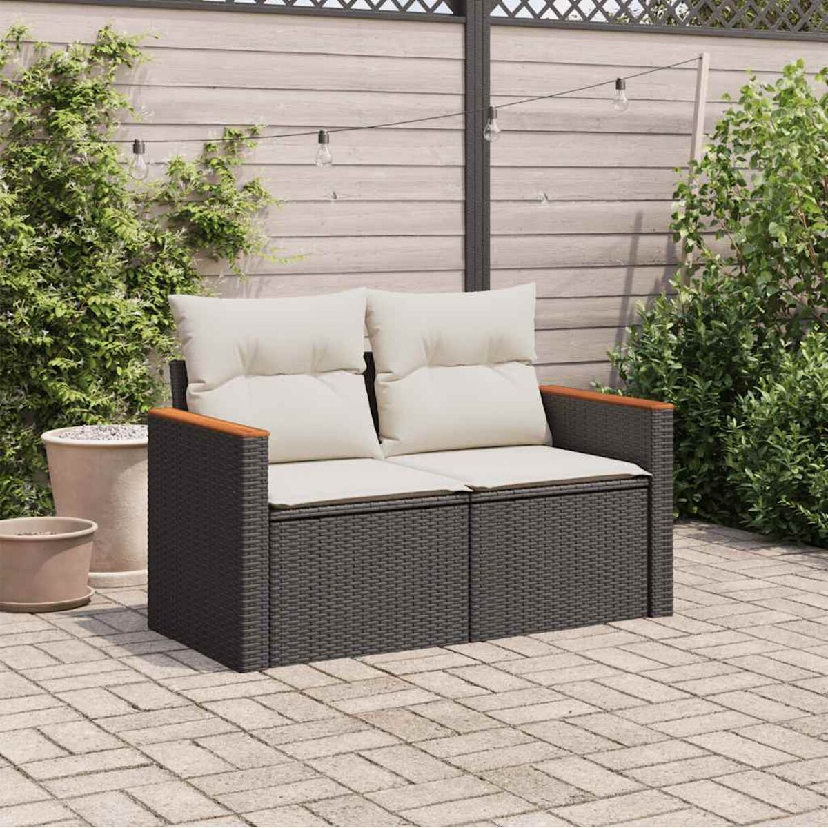 Divano in rattan da esterno con cuscini bianchi. Su un patio in mattoni con piante sullo sfondo.