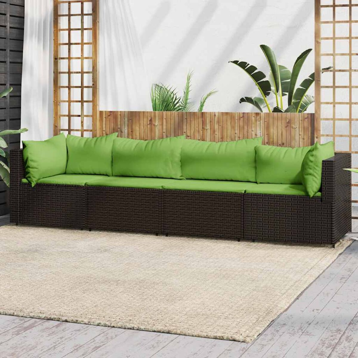 Braunes Korbmöbel-Sofa mit lindgrünen Kissen auf einem beigefarbenen Teppich. Pflanzen dahinter auf Holzterrasse.
