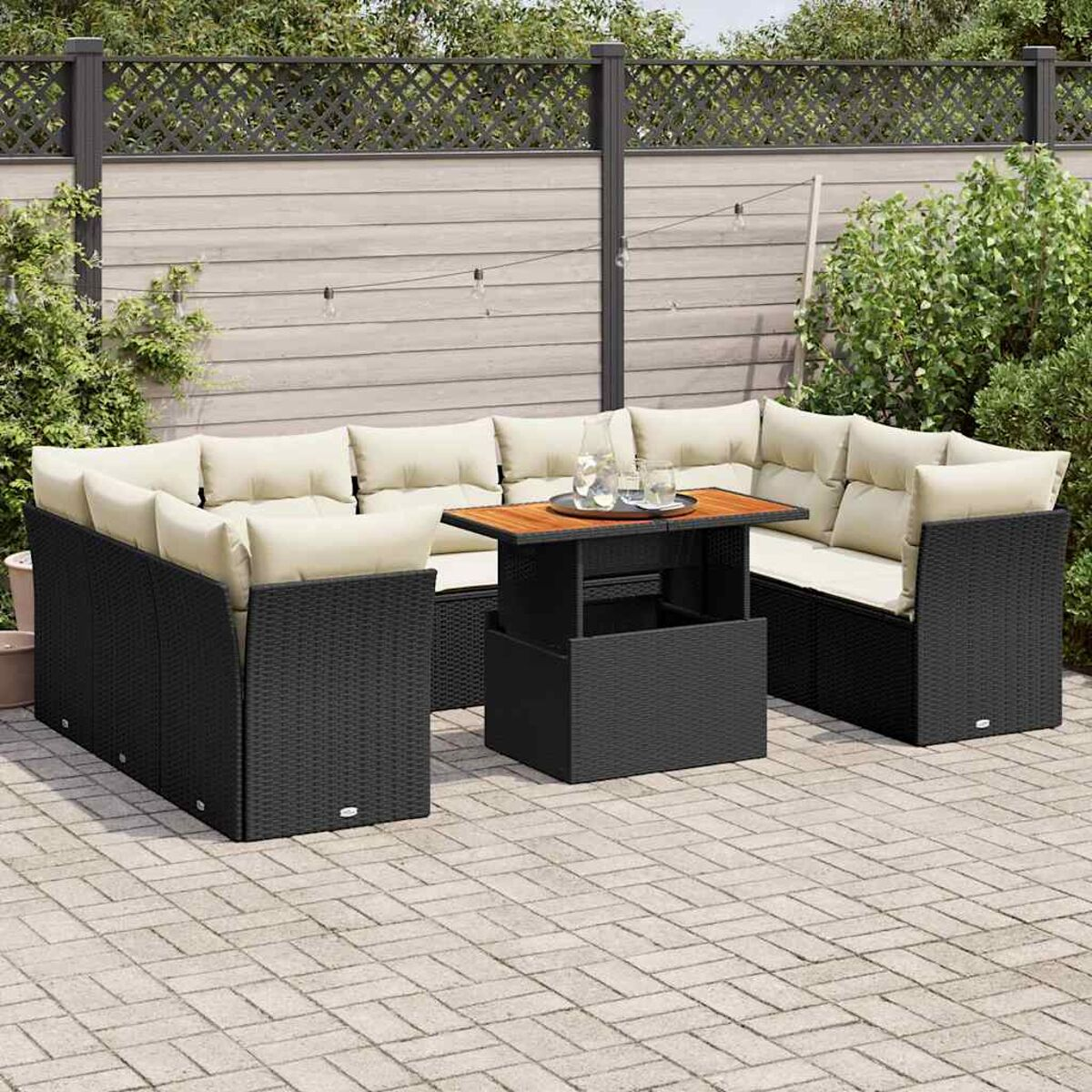 Set di sedute da esterno in vimini nero con tavolo. Ha cuscini color crema. Posizionato su un patio con una recinzione sullo sfondo.