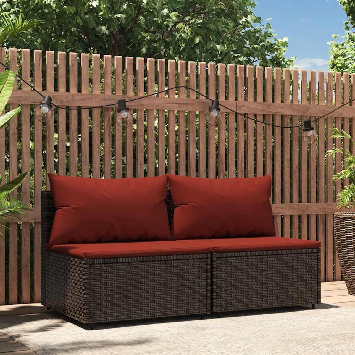 Braunes Korbmöbel-Sofa im Freien mit roten Kissen. Holzzaun-Kulisse und Lichterkette.