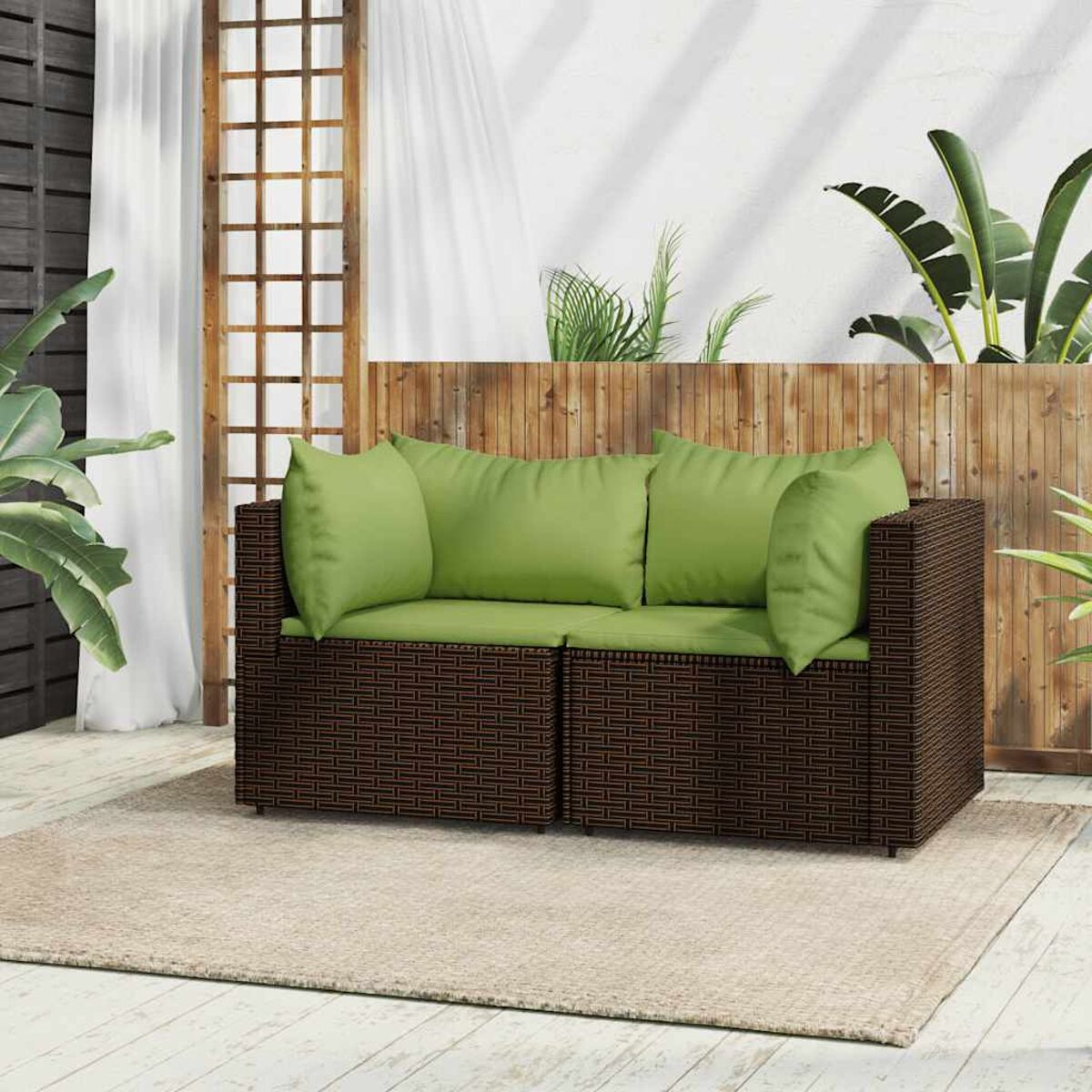 Ein braunes, gewebtes Outdoor-Sofa mit grünen Kissen, auf einem beigen Teppich vor einem Holzzaun.