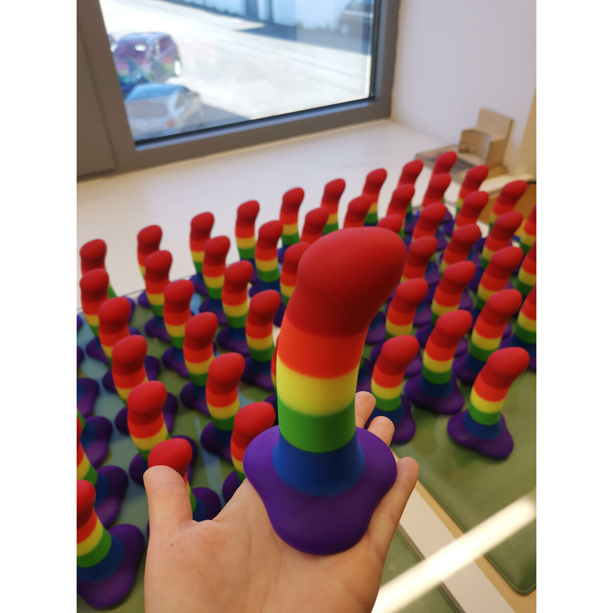 Ręka trzymająca tęczową zabawkę erotyczną, otoczona podobnymi zabawkami. Tłem jest okno.