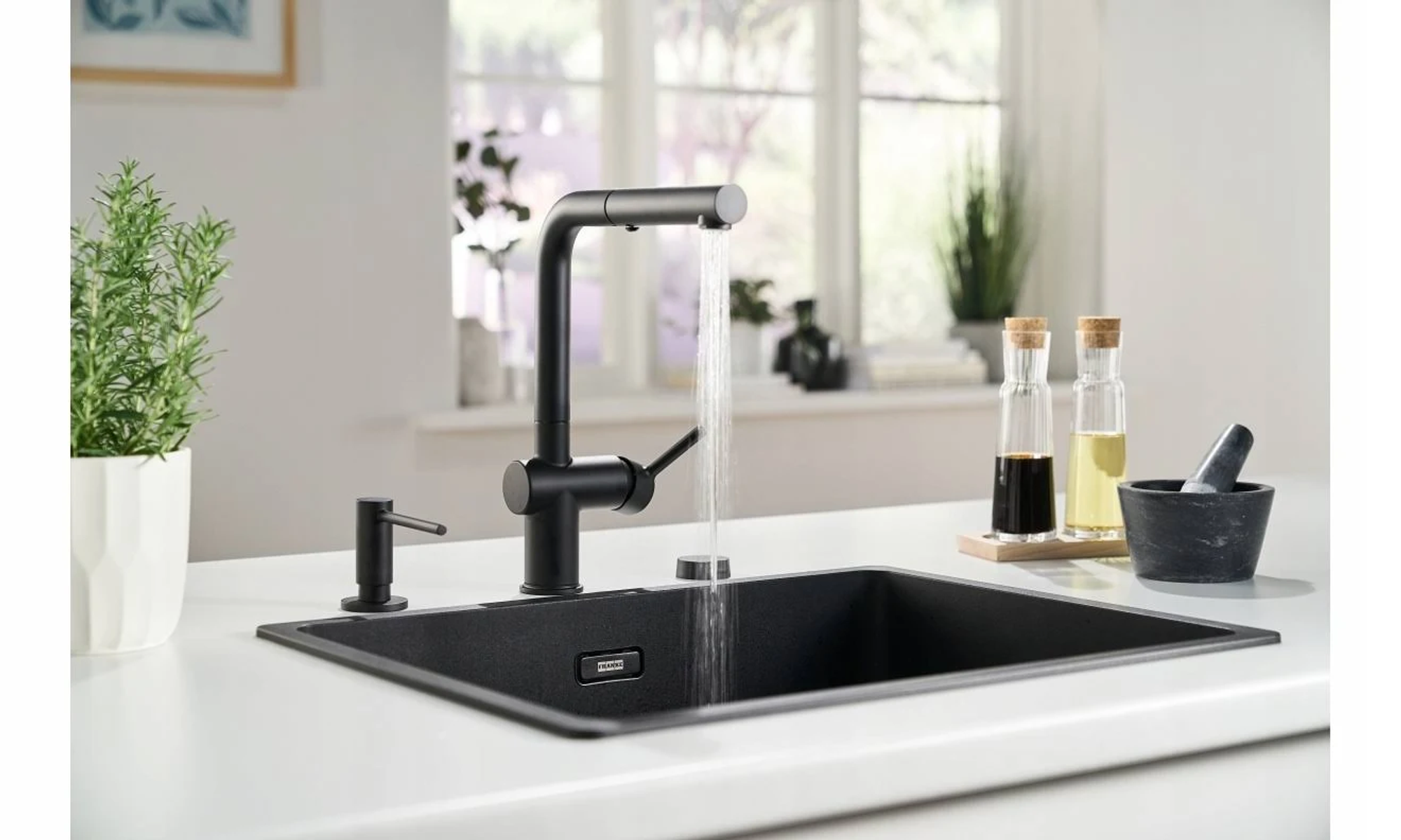 Rubinetto da cucina nero con acqua che scorre in un lavandino nero. Dispenser di sapone, bottiglie.