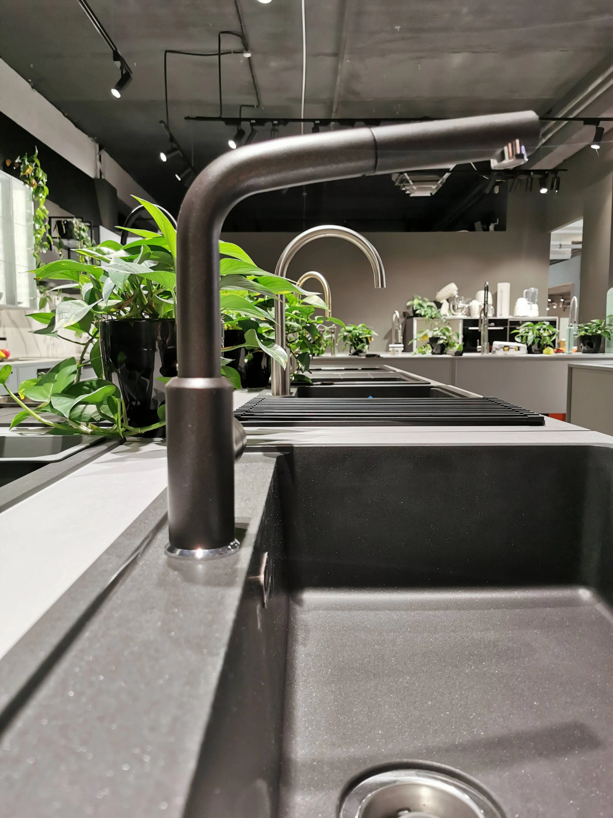 Molteplici lavelli e rubinetti da cucina in uno showroom. Piante e luci sullo sfondo, grigio scuro.