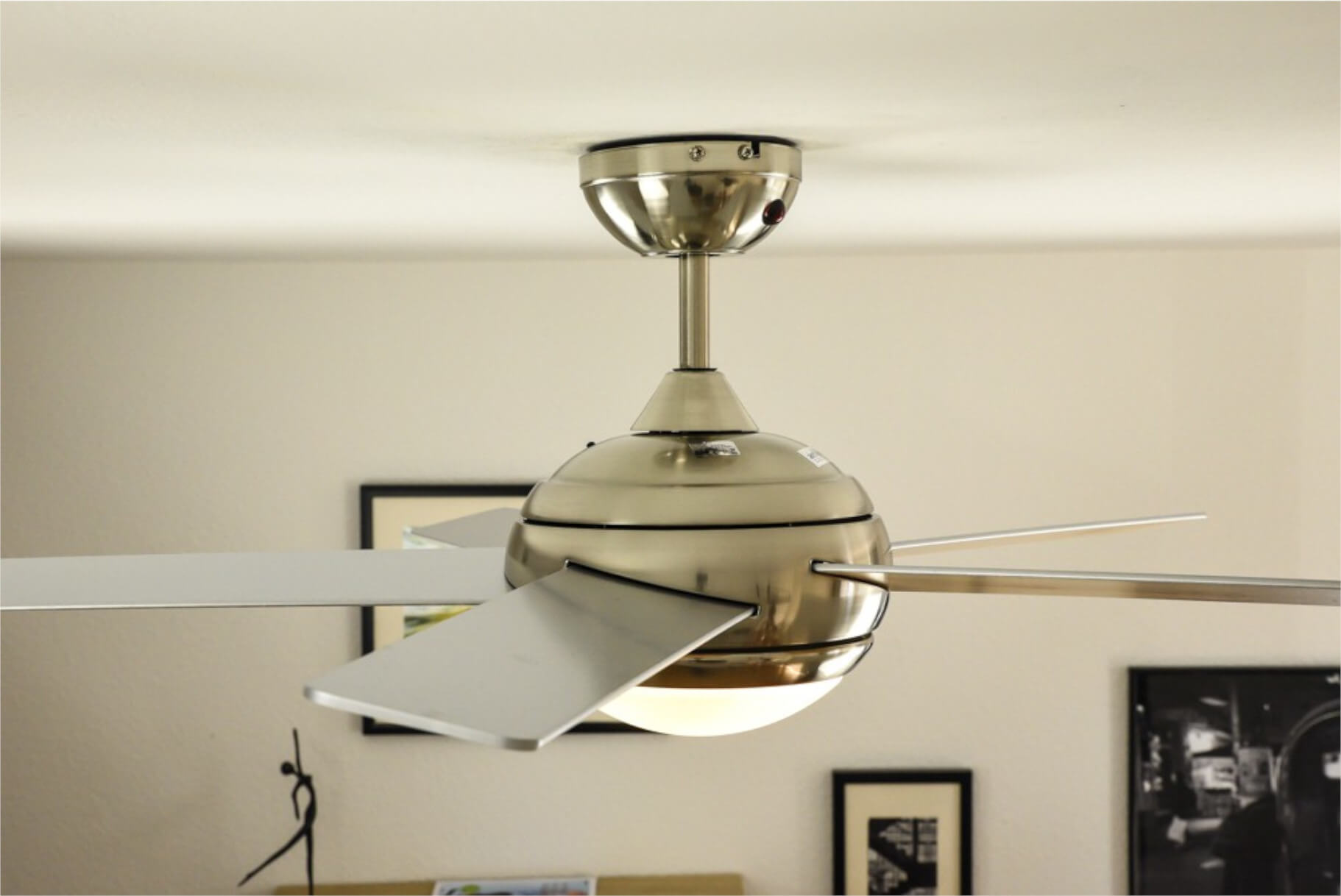 Deckenventilator mit Lichtern, silberne Oberfläche. Bilder im Hintergrund. Neutrale Wände.