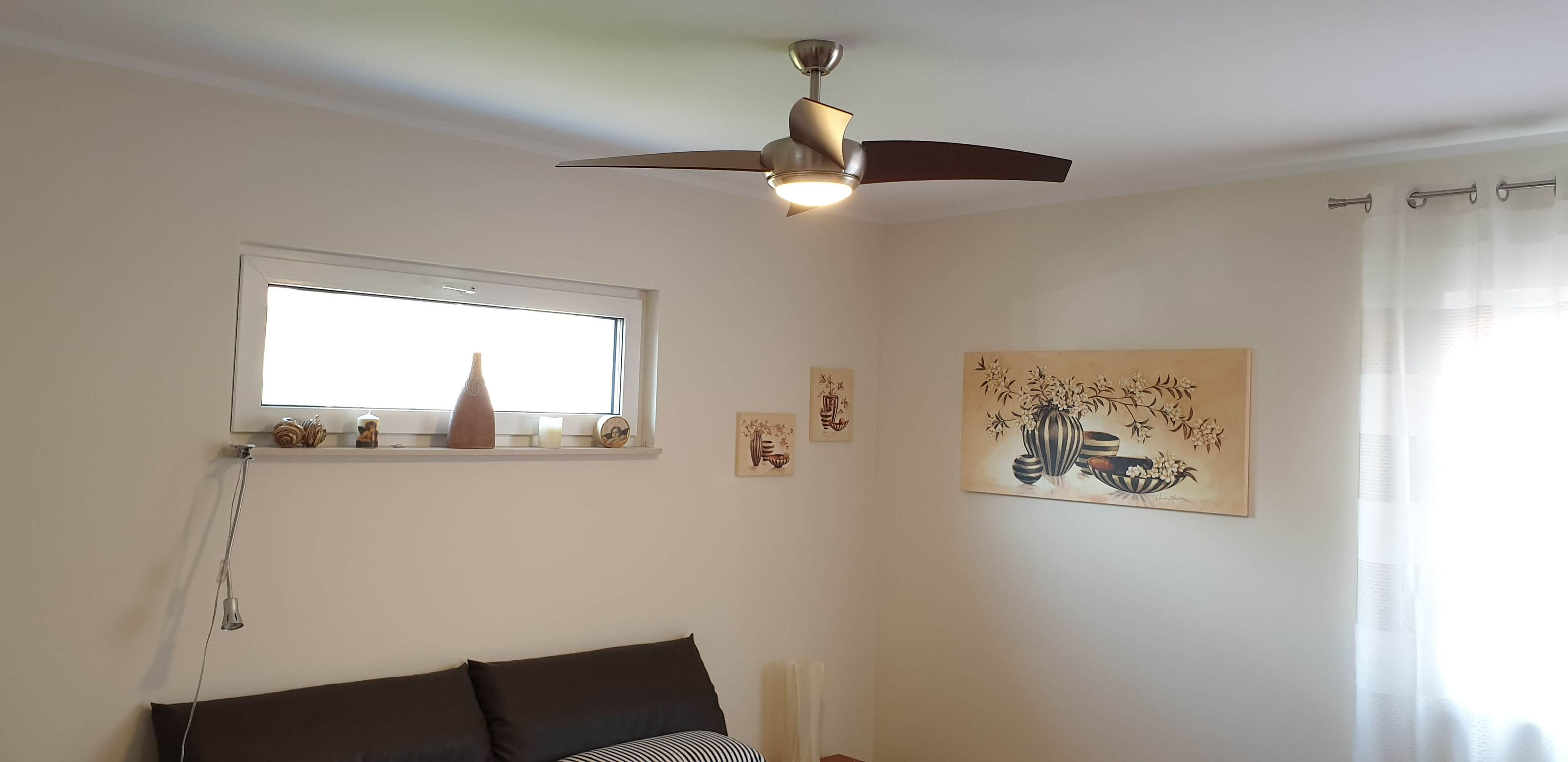 Ein Rauminnenraum. Der Raum hat einen Deckenventilator, ein Fenster, ein Gemälde, ein Sofa und eine Lampe.