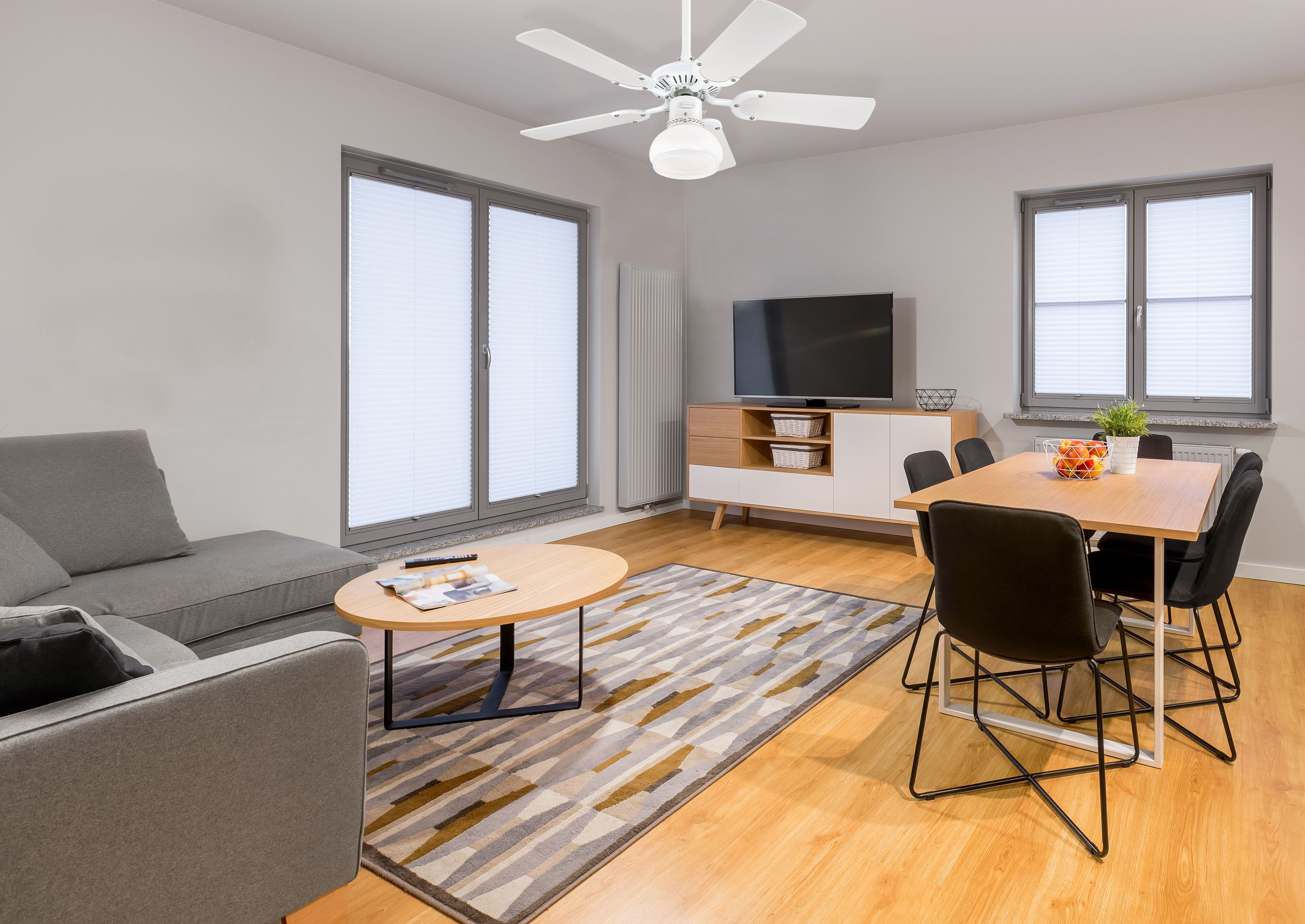 Ein Wohnzimmer mit grauem Sofa, Teppich und Deckenventilator. Fenster und Essbereich sind sichtbar.
