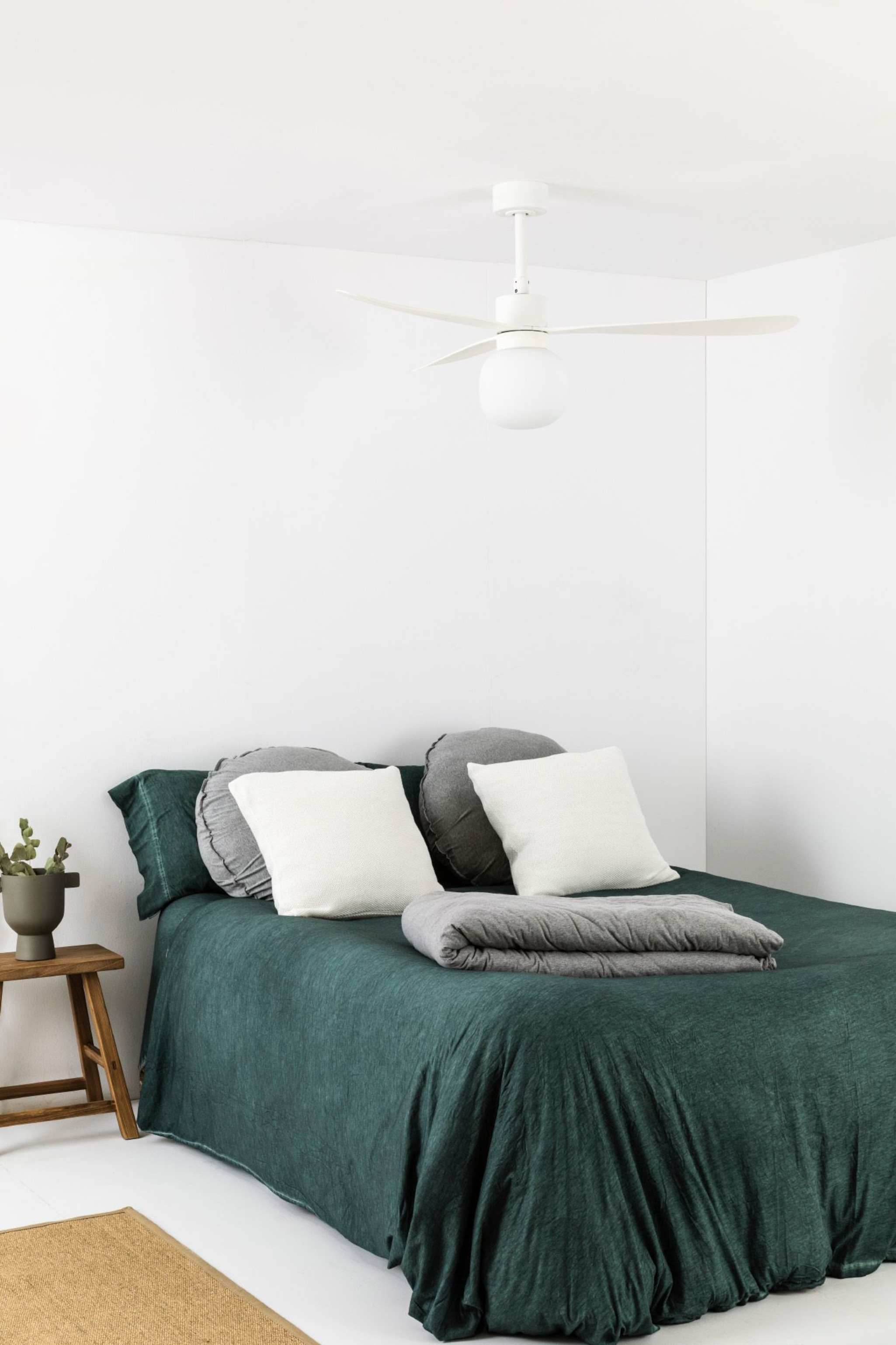 Ein Schlafzimmer mit grüner Bettdecke, Kissen und Deckenventilator. Weiße Wände und Boden.