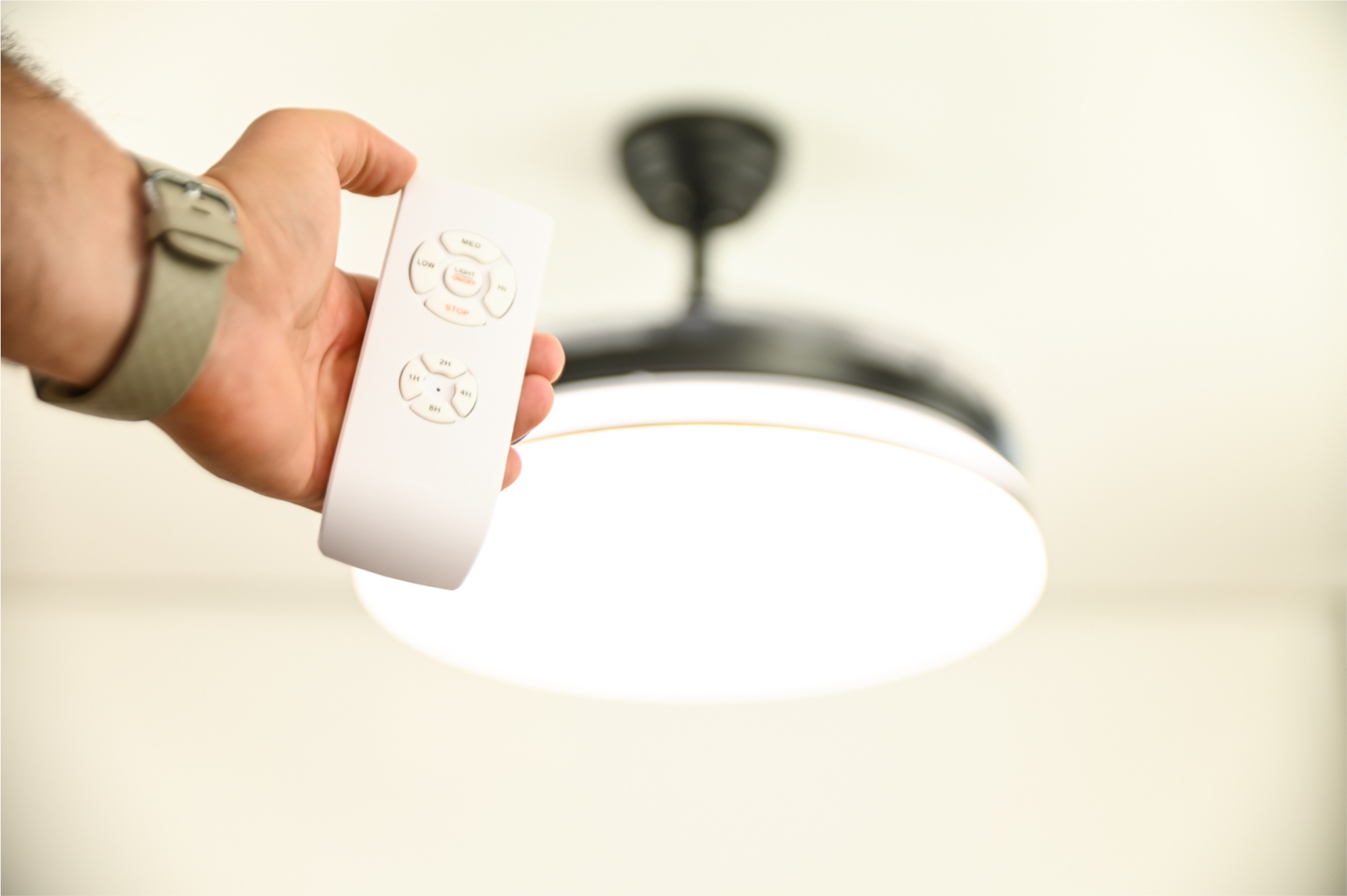 Eine Hand, die eine weiße Fernbedienung hält und auf einen Deckenventilator mit Licht zeigt.