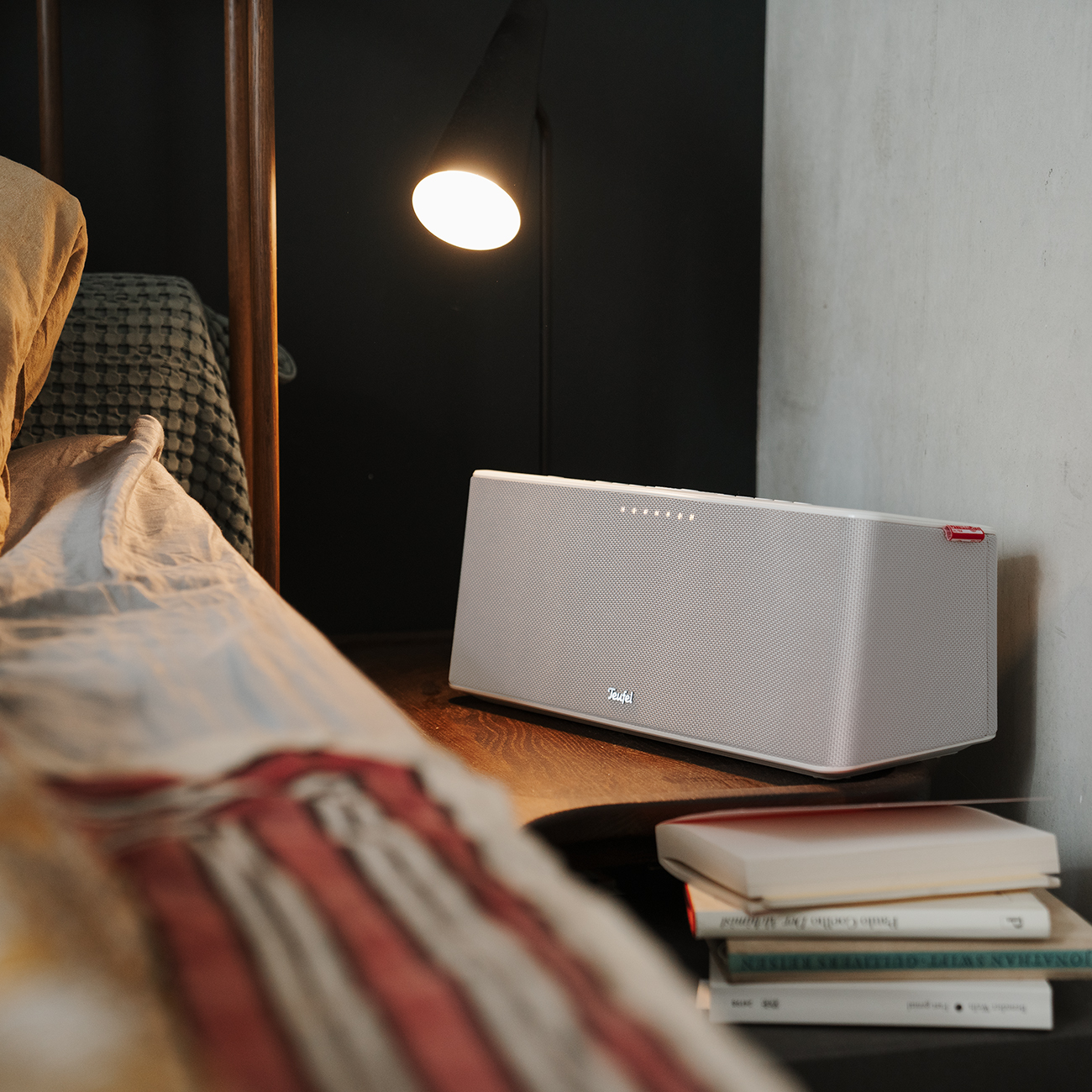 Een witte luidspreker op een nachtkastje met boeken en een lamp. Het bed is zichtbaar.