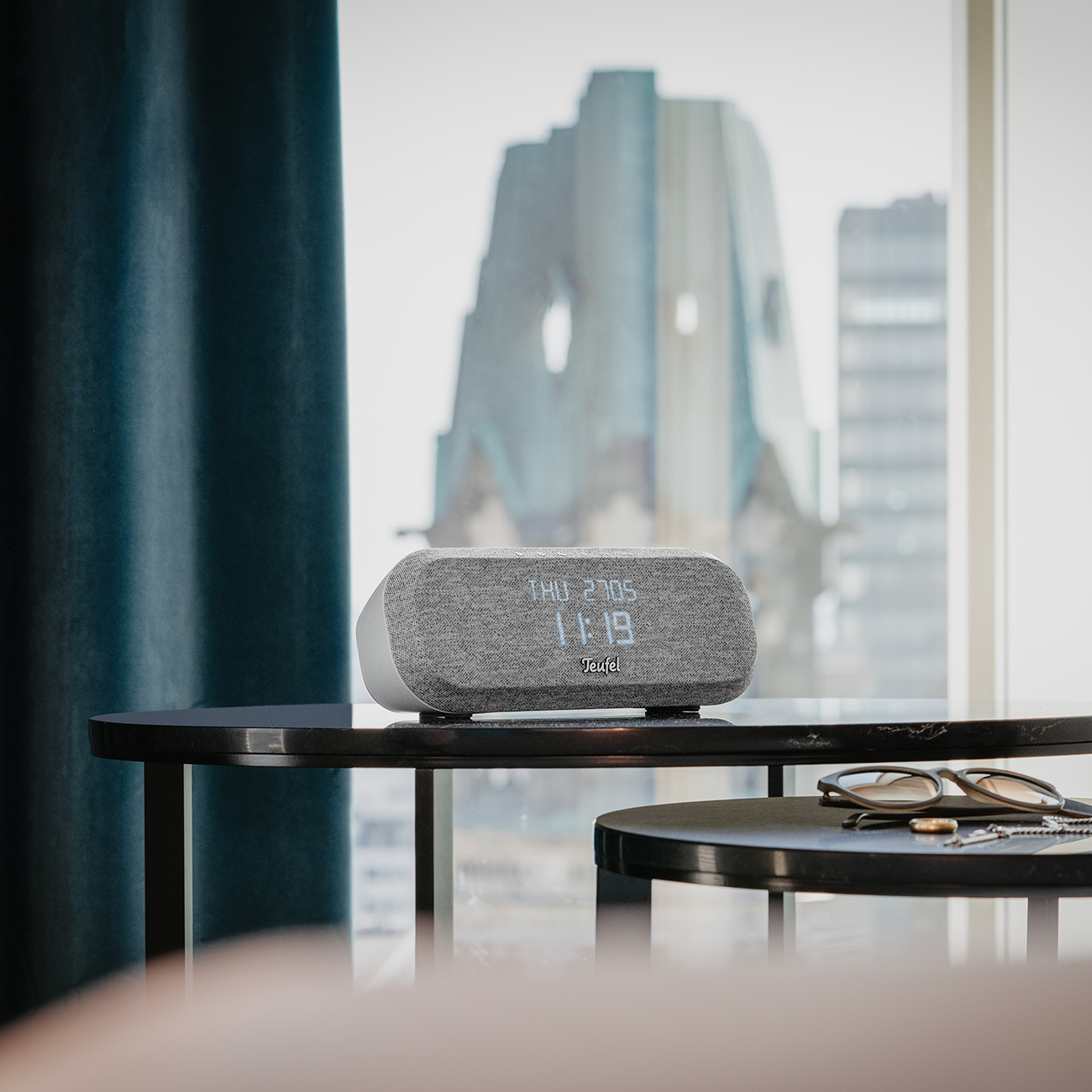 A gray clock radio sits on a table. A window with a city view is in the background.