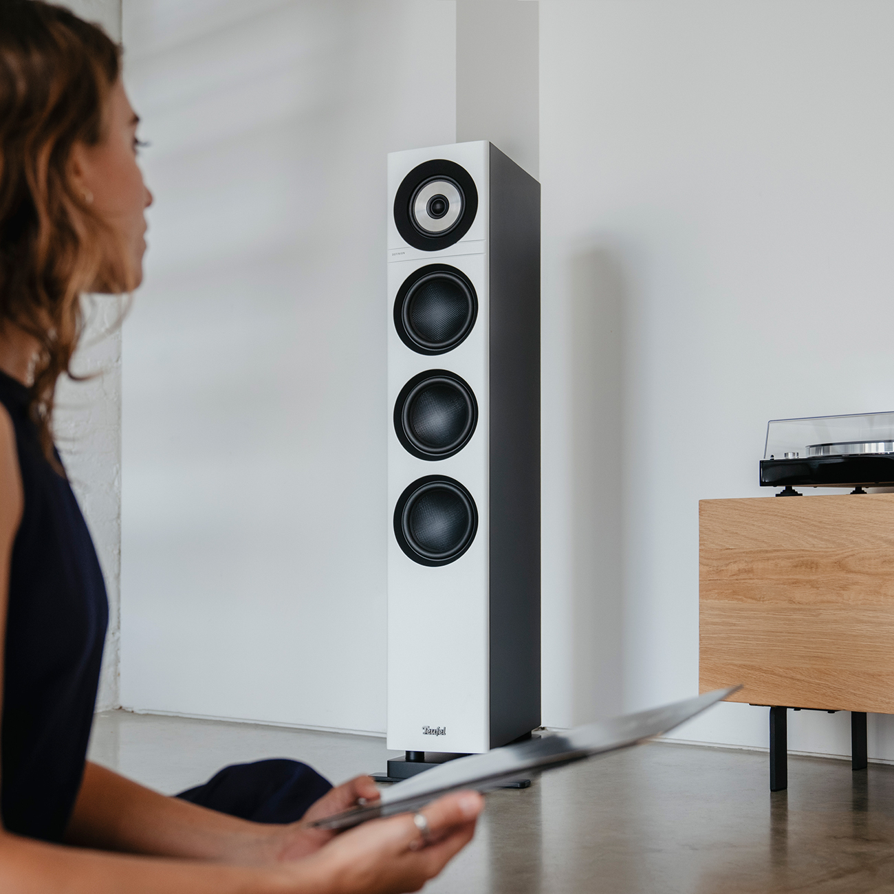 Vrouw met plaat, witte speaker, platenspeler en licht houten kast tegen witte muur.