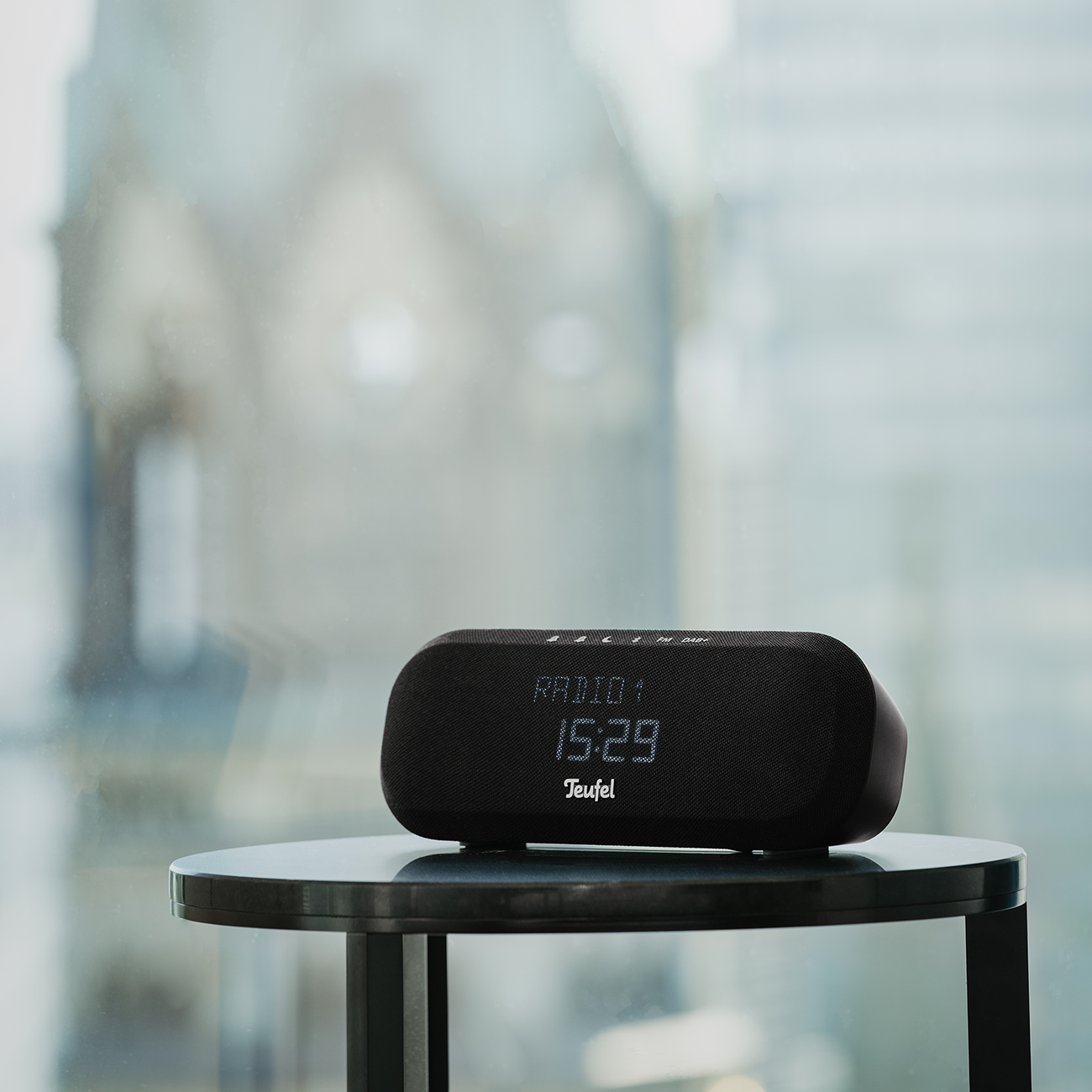 Black Teufel digital clock radio on a table, with a blurred window background.