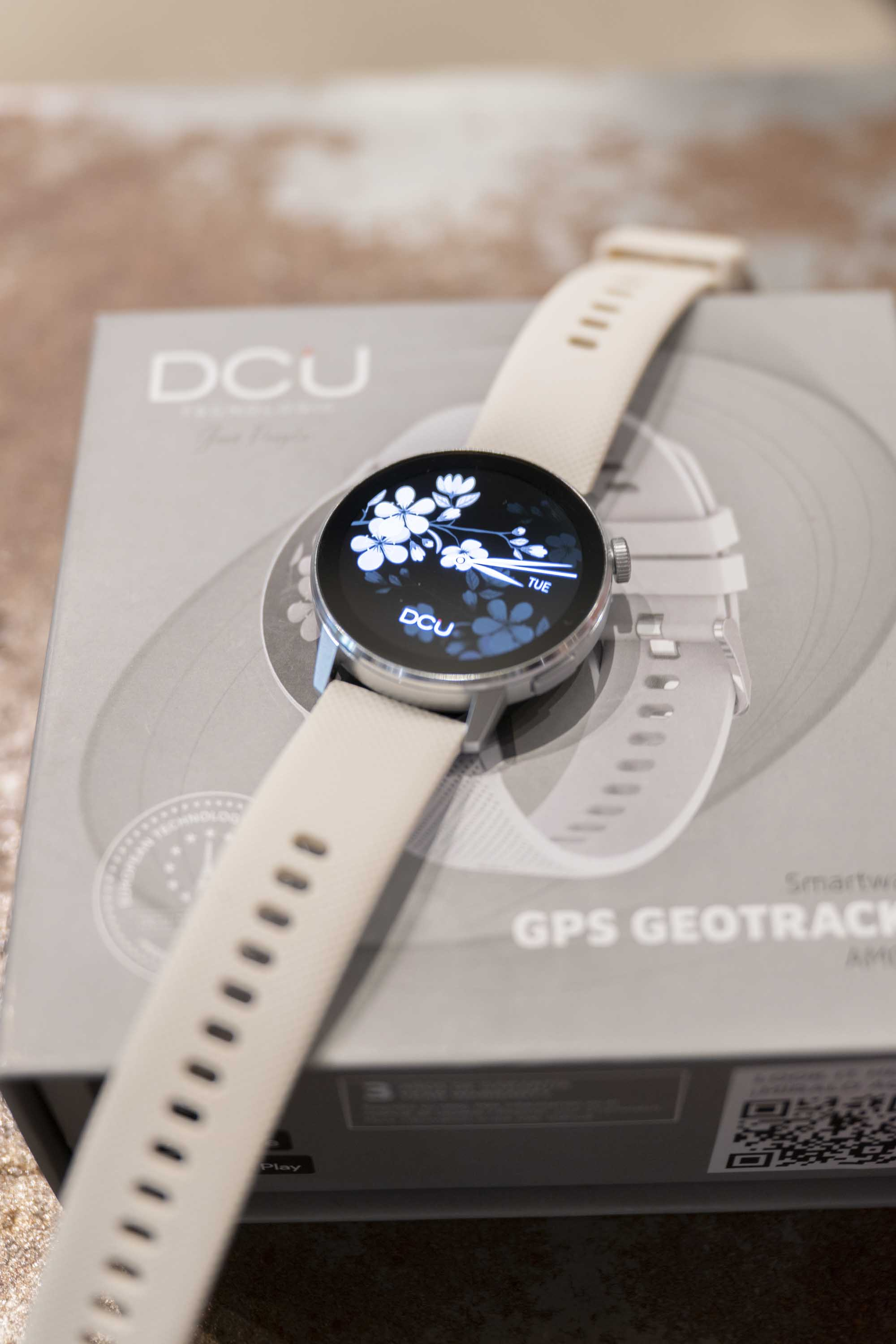 Smartwatch con pantalla floral sobre una caja. Correa beige, caja gris con texto. Iluminación suave.