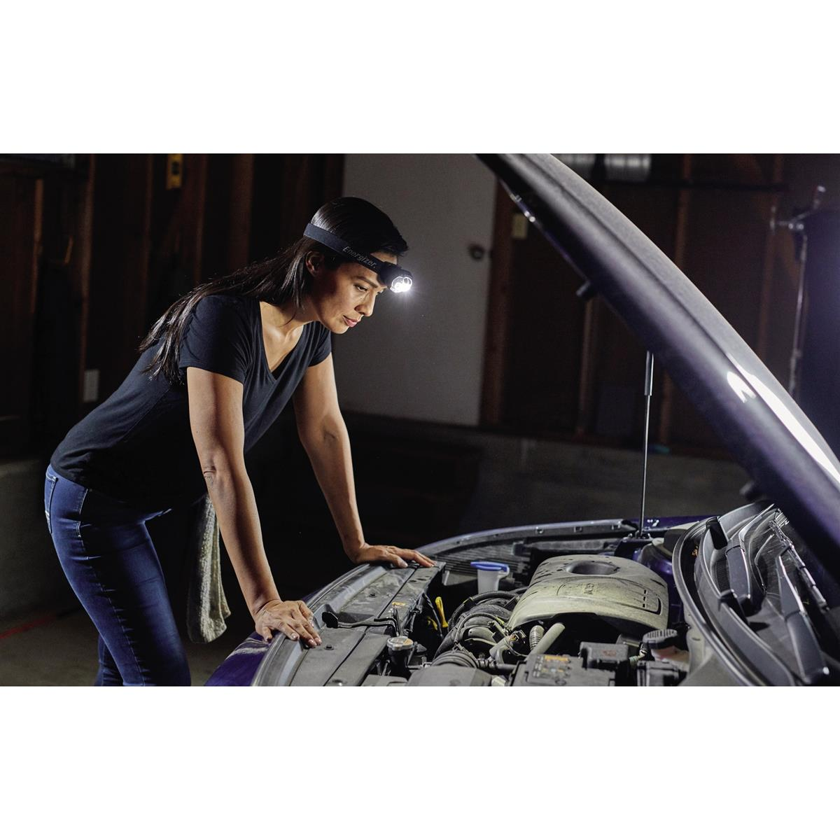 Frau mit Stirnlampe untersucht einen Automotor. Garage mit offener Motorhaube.