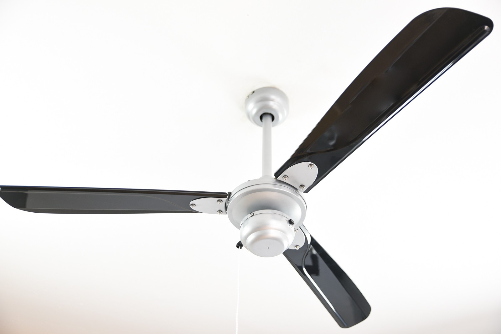 Un ventilatore da soffitto con tre pale nere e un motore centrale argentato, visto dal basso contro un soffitto bianco.