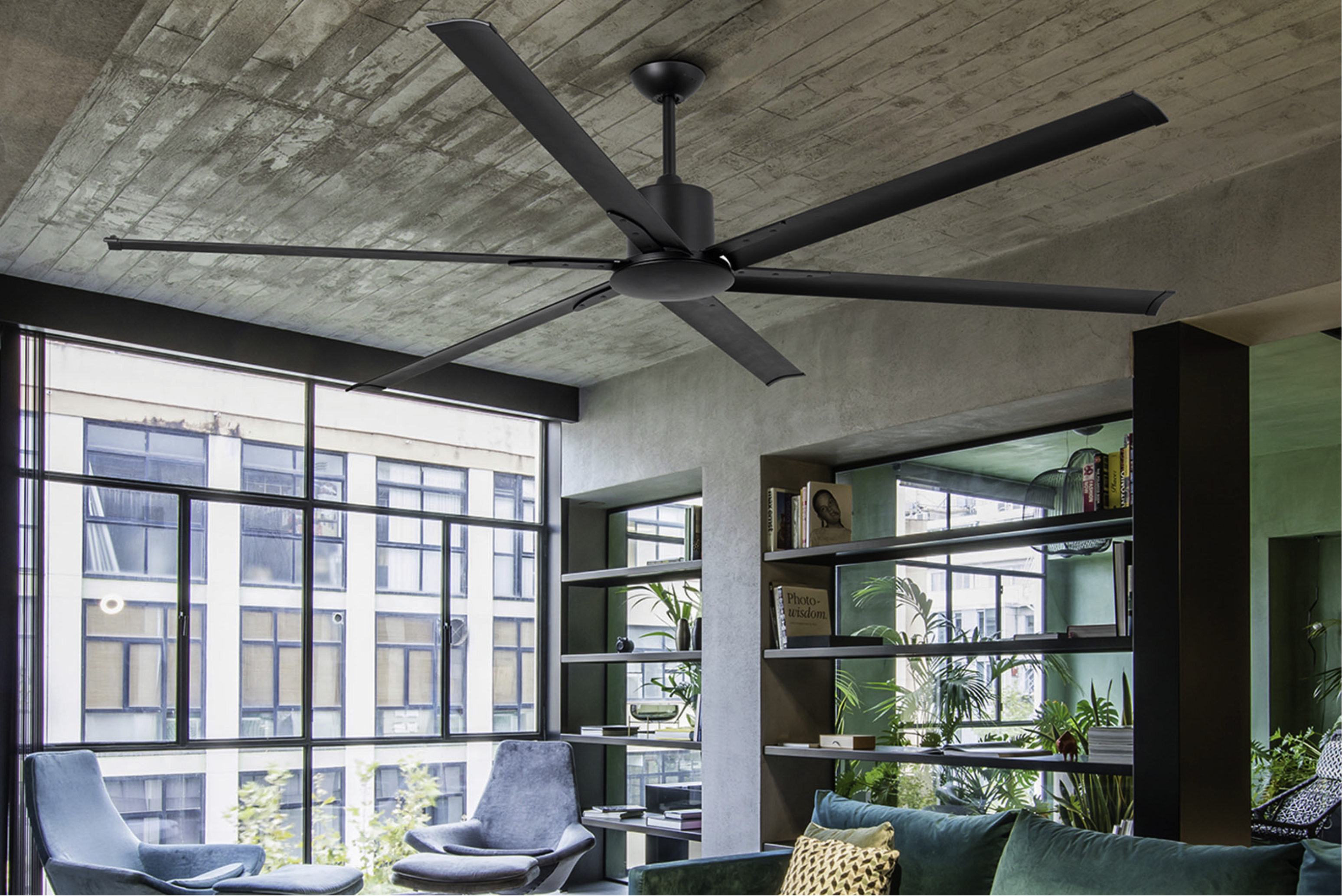 Un grande ventilatore da soffitto nero con sei pale, in una stanza. Soffitto in cemento.