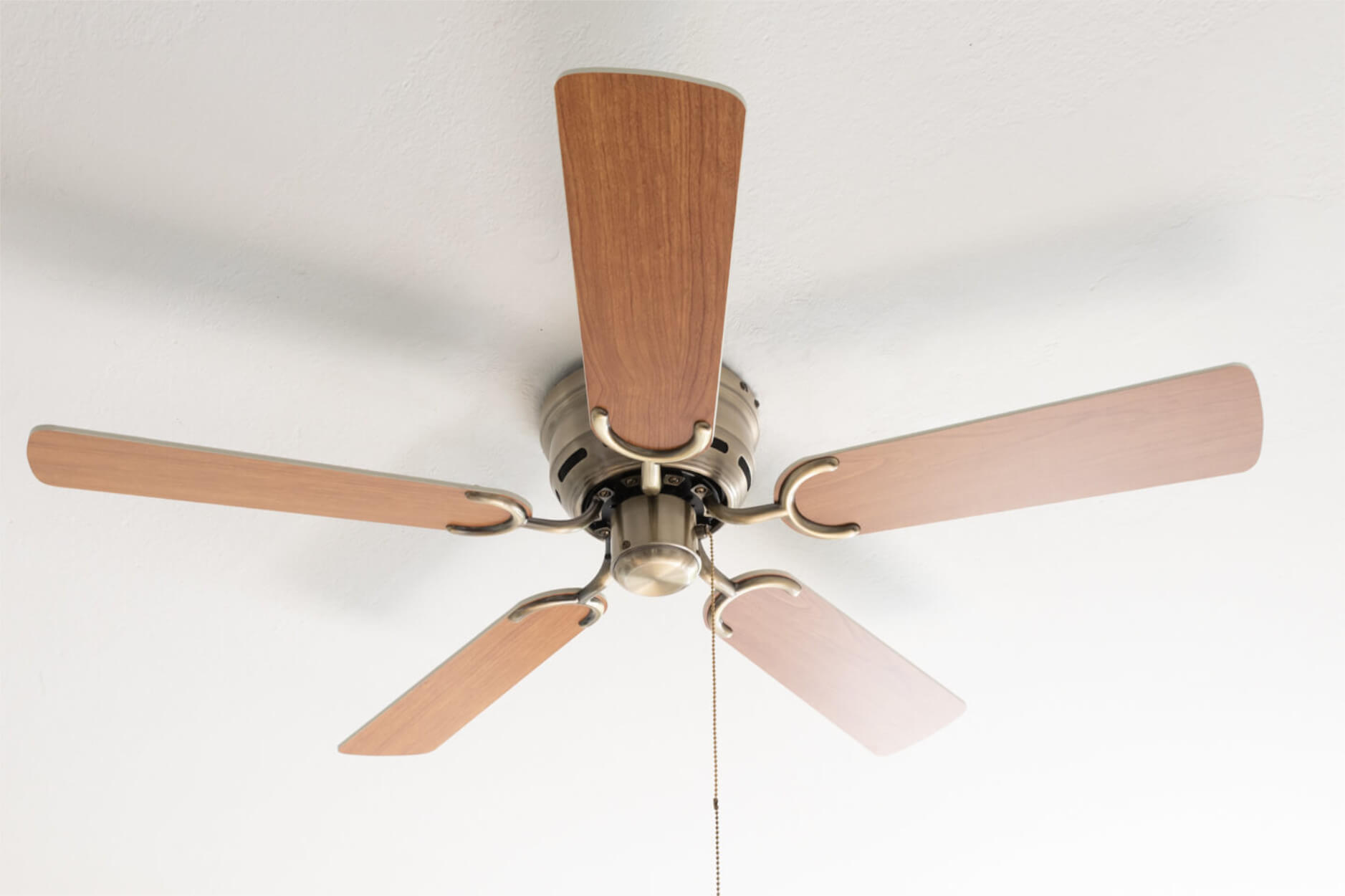 Ein Deckenventilator mit fünf Holzflügeln und einem silbernen Sockel, vor einer weißen Decke.