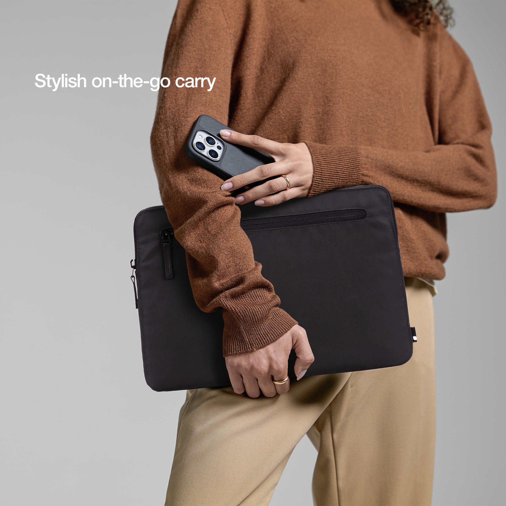 Donna che tiene una custodia per laptop nera e un telefono, indossa un maglione marrone, sfondo neutro.