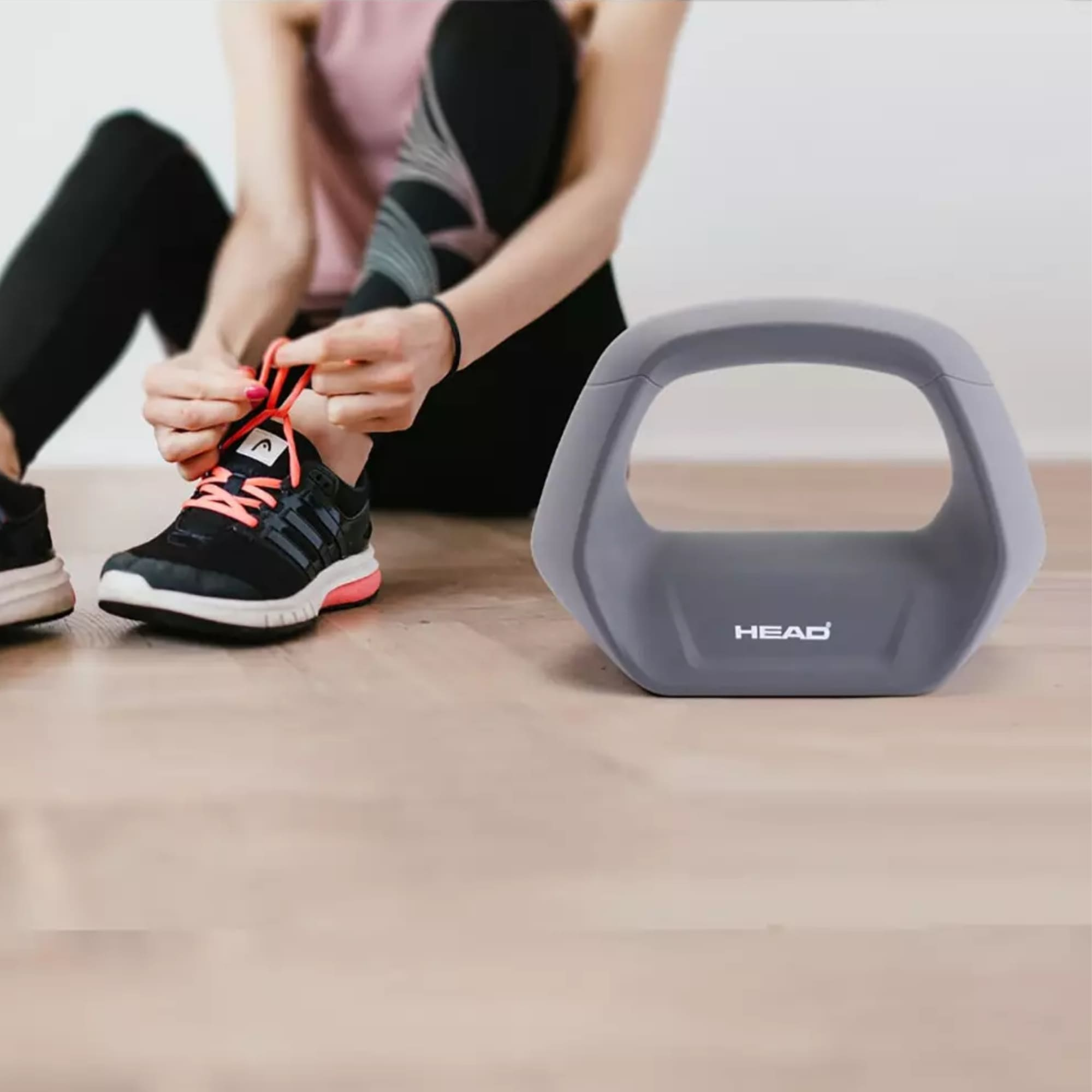 Een vrouw die haar veters strikt naast een grijze HEAD kettlebell op een houten vloer.