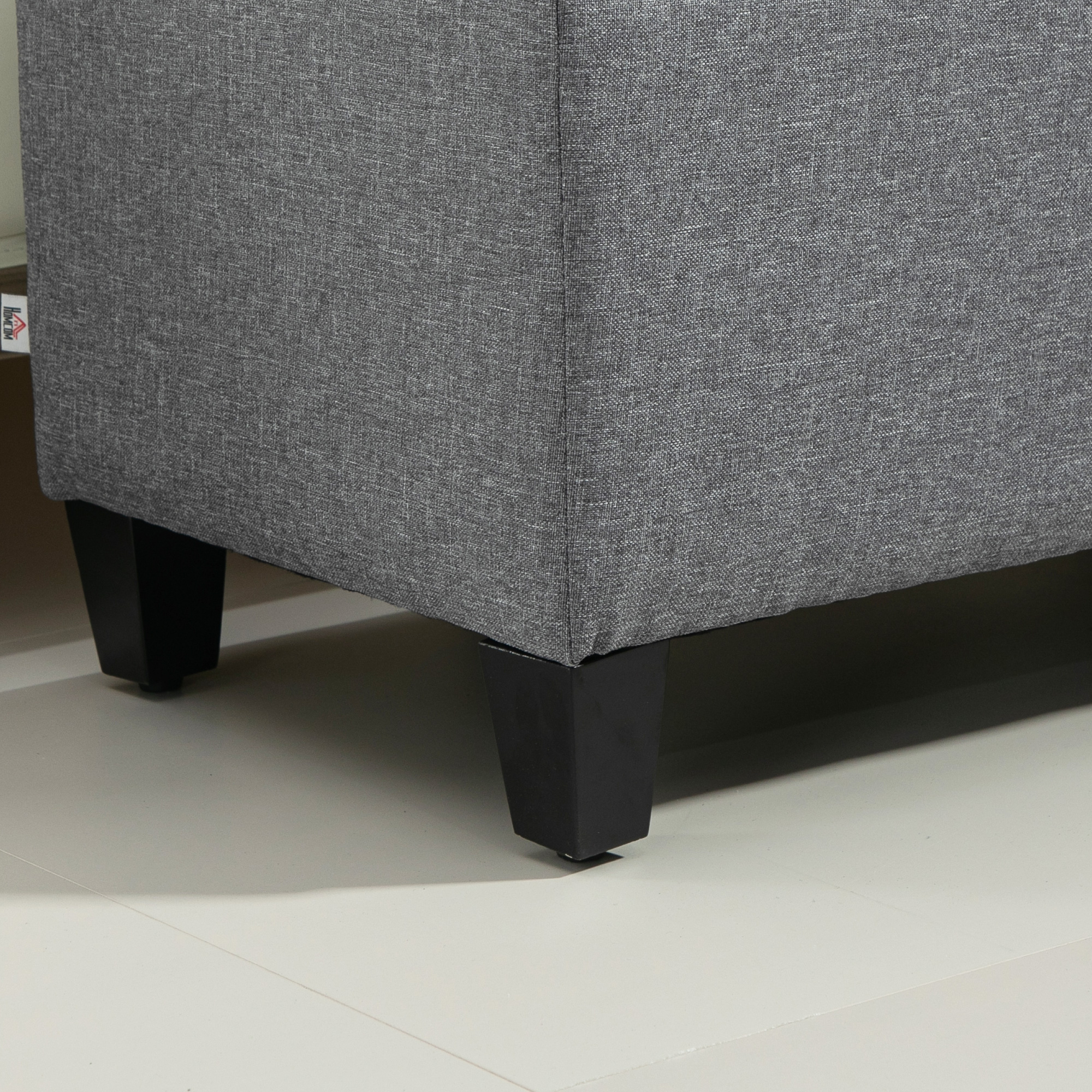 Nahaufnahme einer grauen Stoffcouch. Schwarze Beine sind an den unteren Ecken sichtbar. Weiße Bodenfliesen.