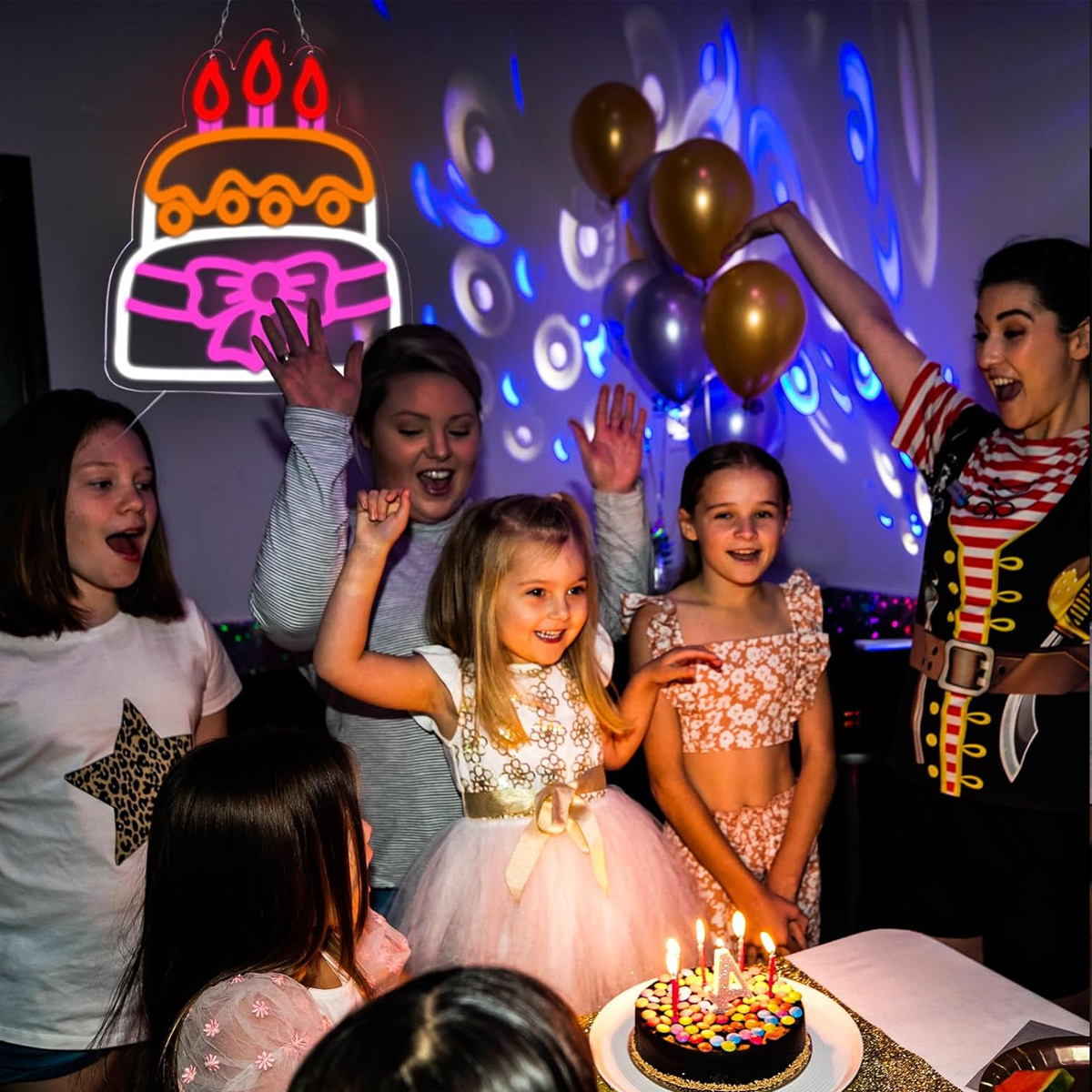 Gruppe von Leuten feiert Geburtstag. Kuchen mit Kerzen, Ballons und einem Neon-Schild.