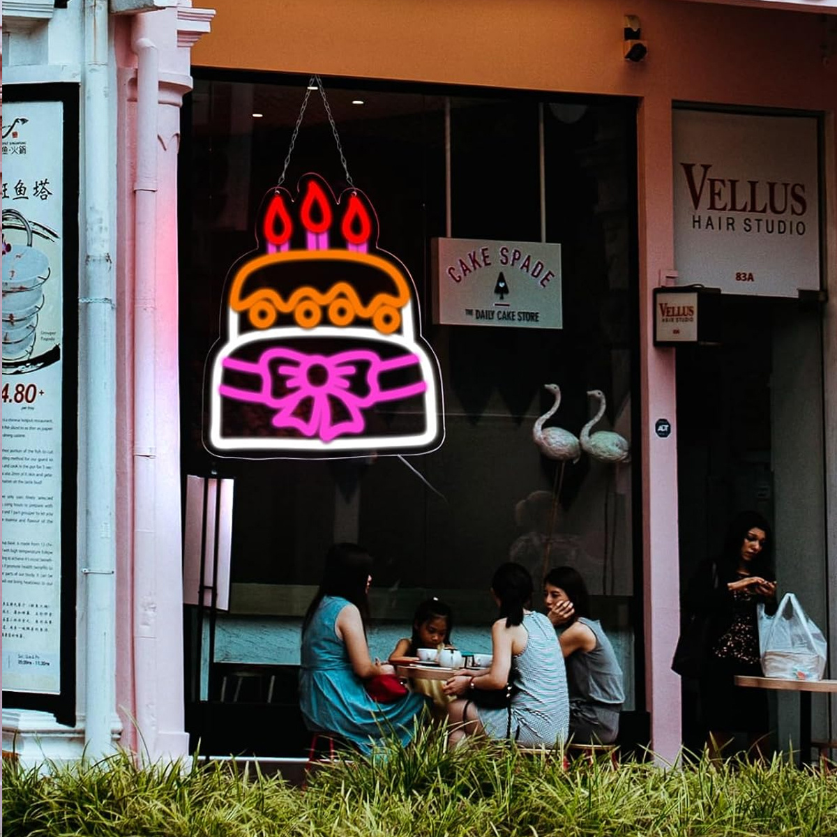 Außenaufnahme eines Cafés mit Neon-Kuchenschild und Leuten an Tischen.