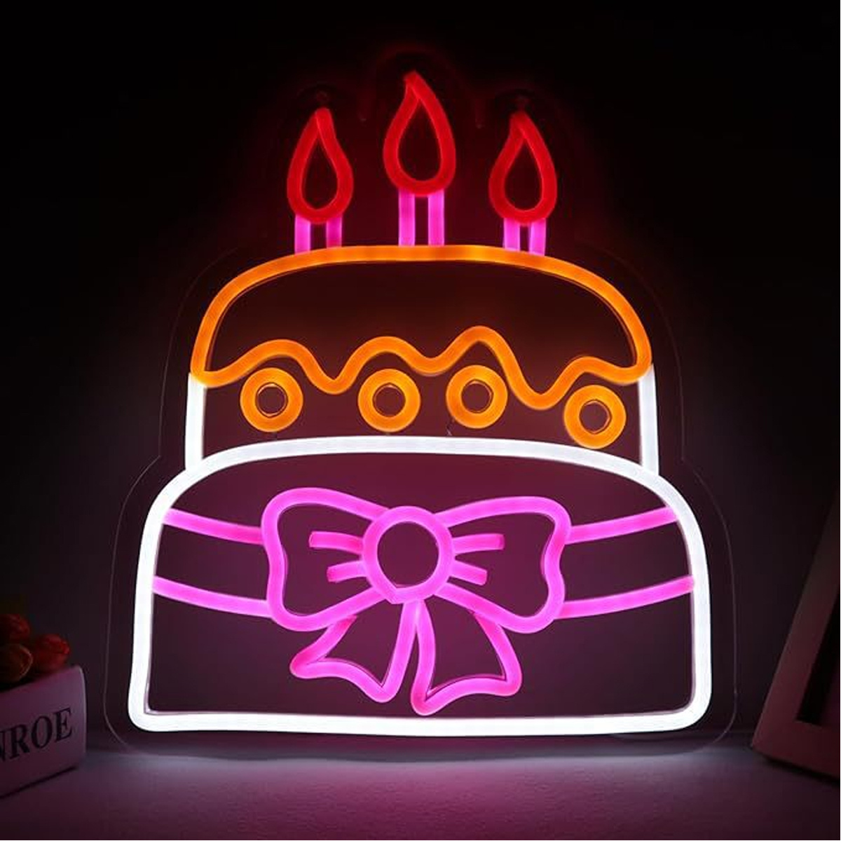 Neon-Kuchenschild mit drei brennenden Kerzen. Rosa Schleife darunter. Schwarzer Hintergrund.