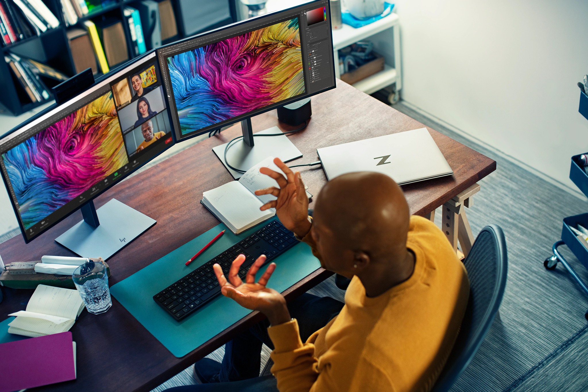 A man is in a meeting on a computer with two monitors. He has his hands up.