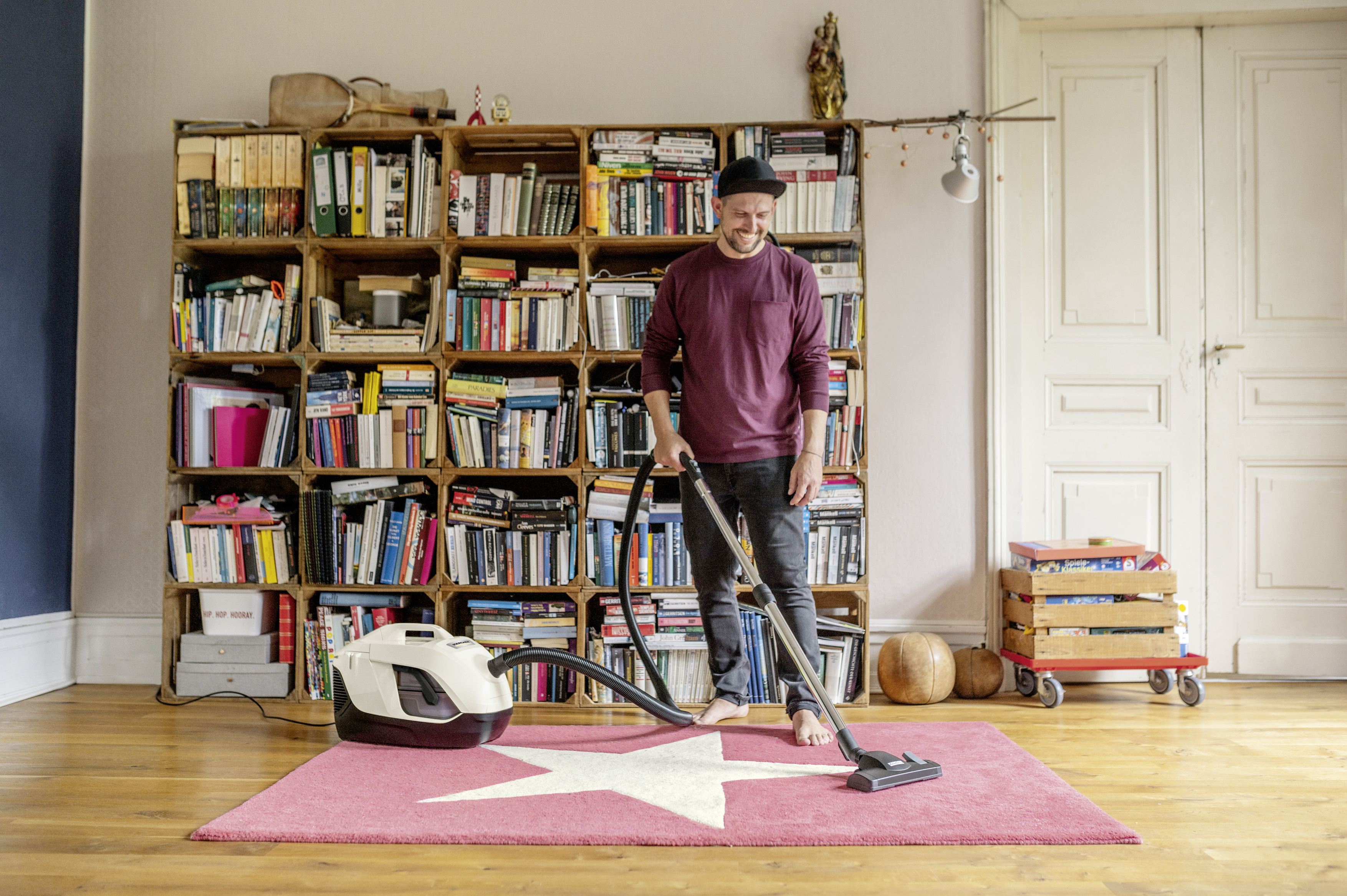 Mężczyzna w bordowej koszuli odkurza różowy dywan z gwiazdą.