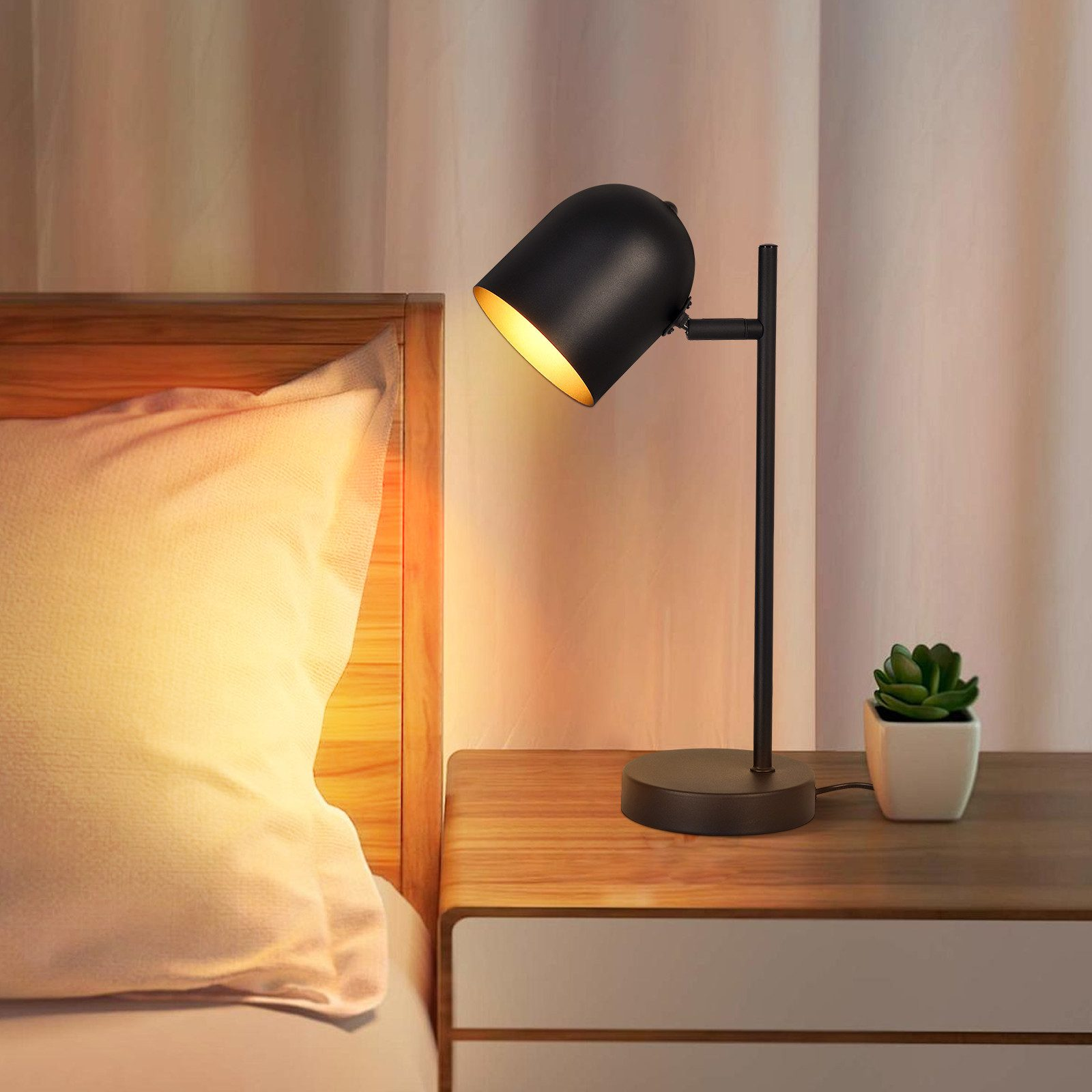 Czarna lampa biurkowa na drewnianej szafce nocnej z małą rośliną. Widoczna biała poduszka i rama łóżka.