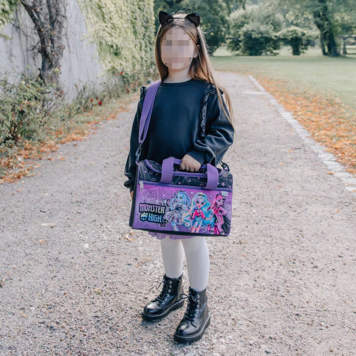 Mädchen in schwarzem Outfit mit Katzenohren, das eine Monster High Tasche hält, geht auf einem Weg.