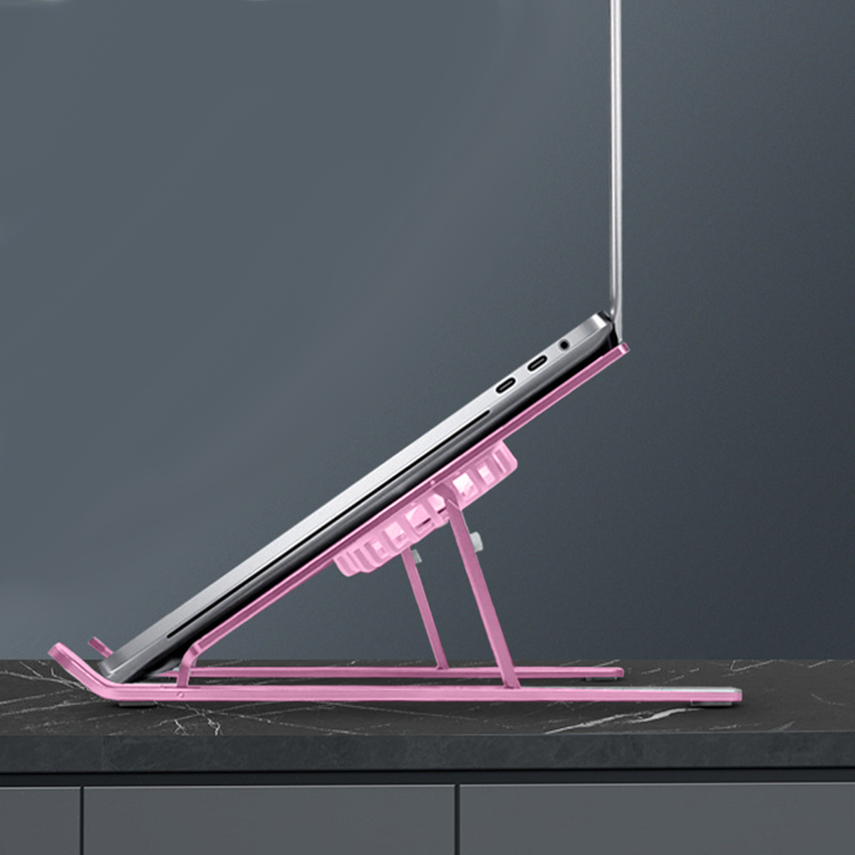 Ein rosa Laptopständer. Ein Laptop ist in einem Winkel auf dem Ständer aufgestellt. Es steht auf dunkler Oberfläche.
