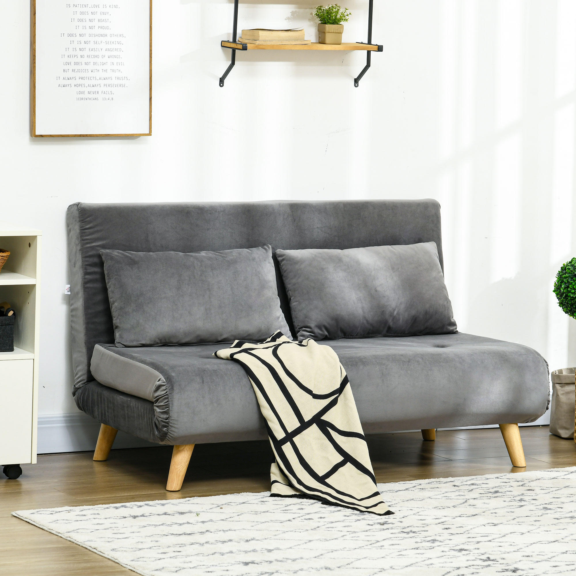 Graues Sofa mit zwei Kissen und einer Decke auf einem Teppich. Wand mit Zitat und Regal mit Pflanzen.