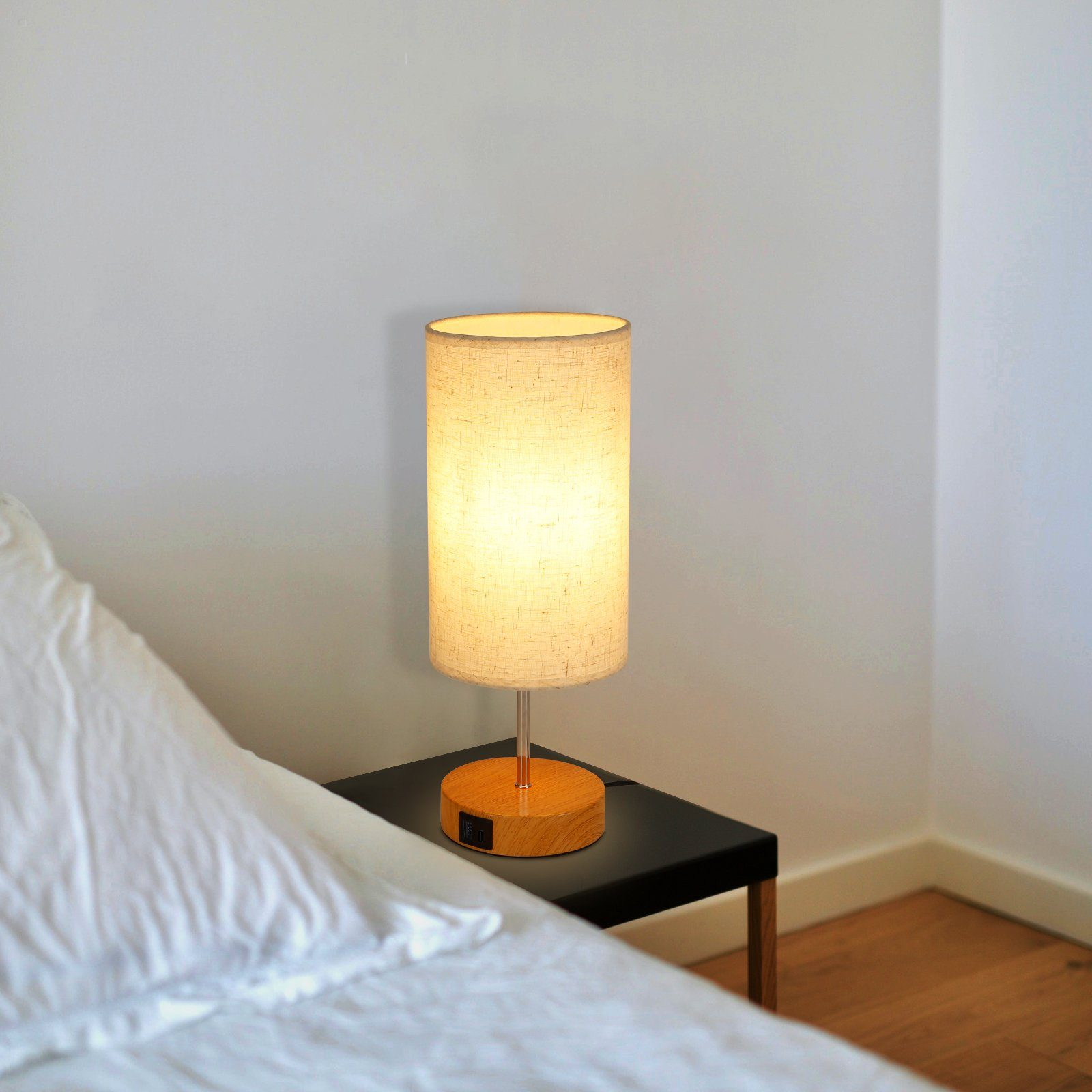 Lampa z beżowym kloszem i drewnianą podstawą na czarnym stoliku obok łóżka.